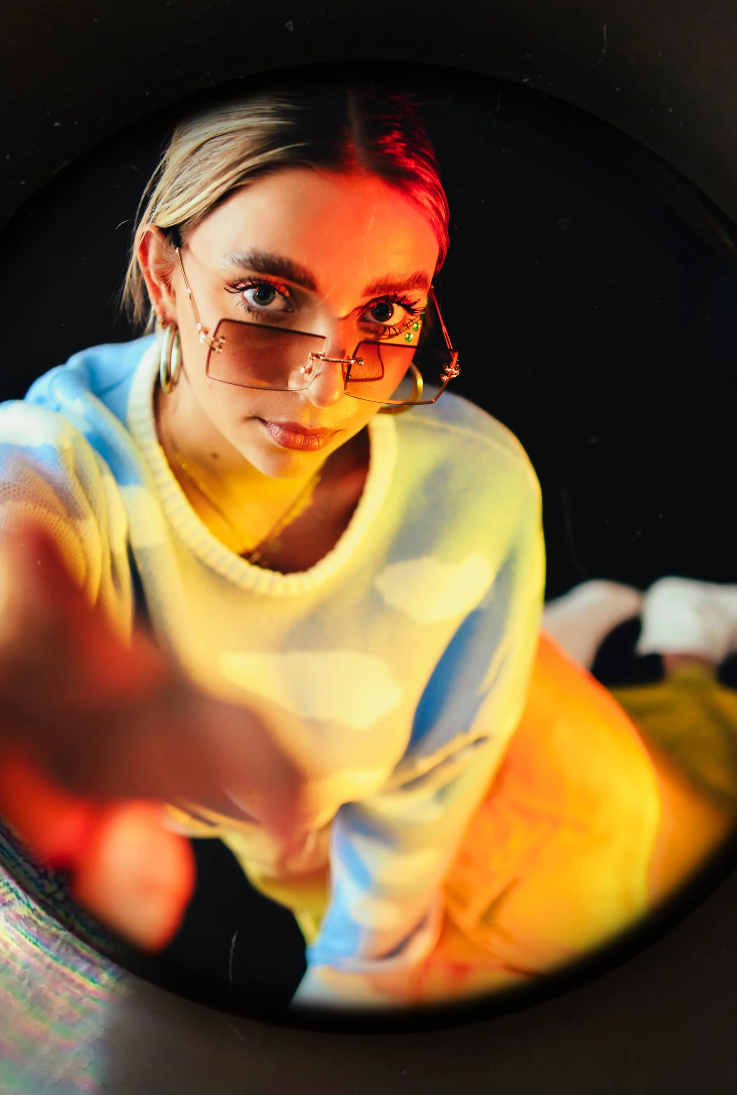 A young woman wearing glasses with a reflection of red light, a pastel-colored sweater, and hoop earrings, taking a selfie with a fisheye lens.
