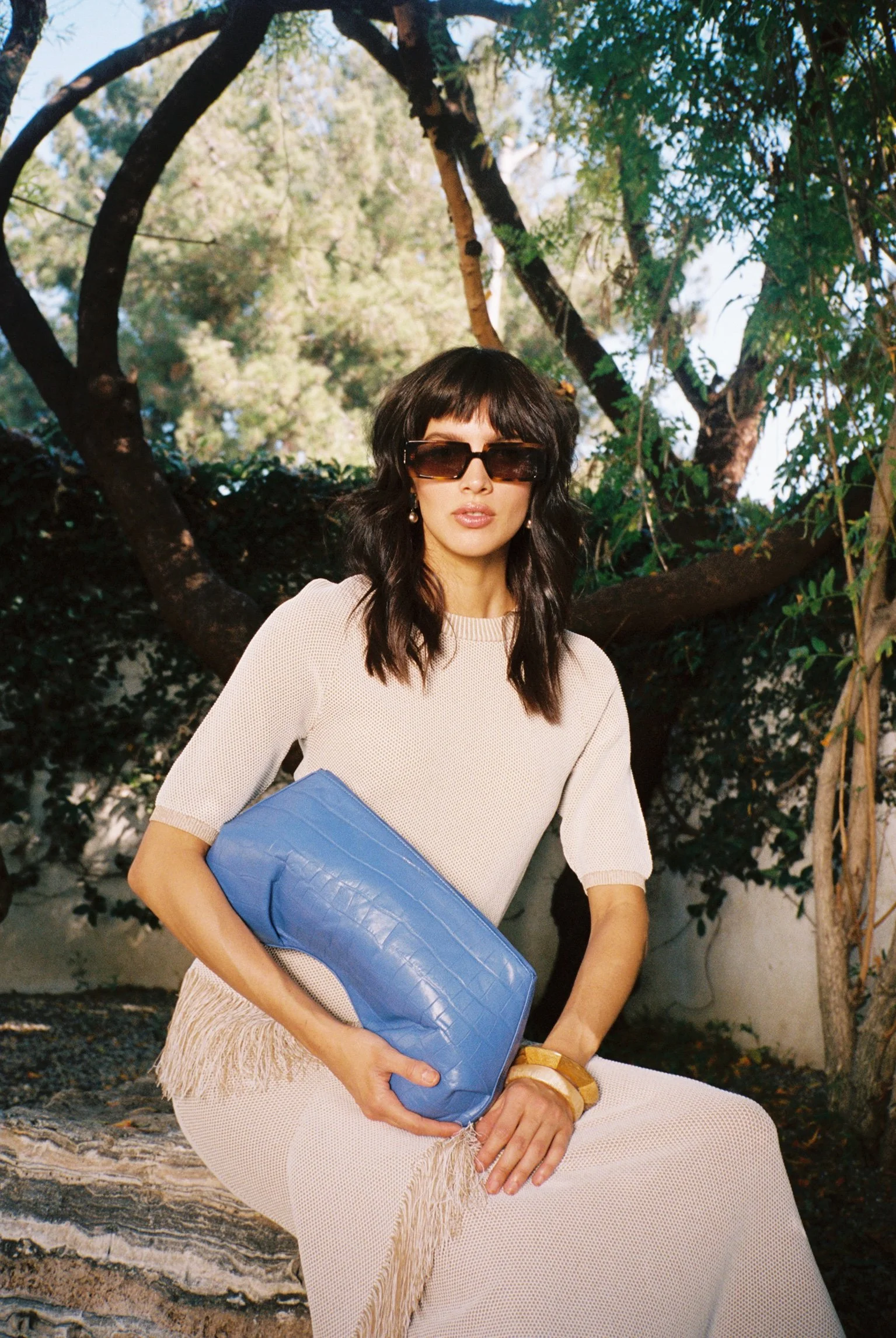 A woman with dark wavy hair wearing large sunglasses, pearl earrings, a beige short-sleeve dress with fringes, and a gold bracelet, sitting outdoors on a log, holding a blue clutch purse, with trees and blue sky in the background.