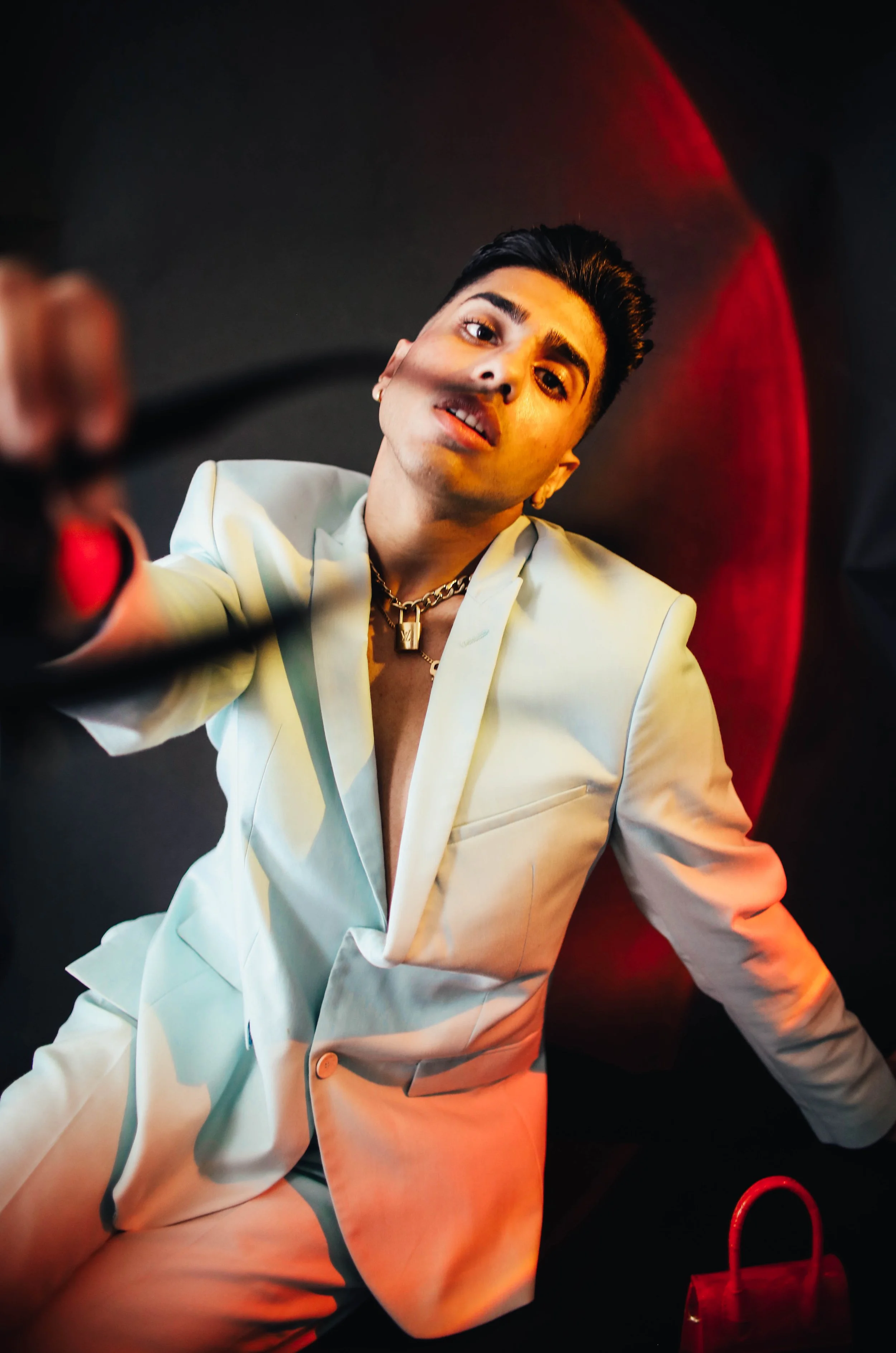 A person in a light-colored suit with a chain necklace, leaning back on a red chair, looking into the camera with a confident expression.