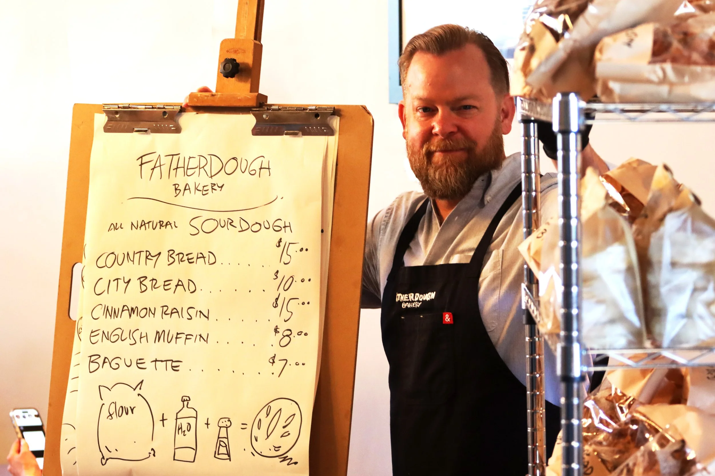 A man with a beard stands behind a bakery menu board, which lists various types of sourdough and country bread. The man appears to be working at Father Dough Bakery, with a rack of wrapped baked goods visible on the right.