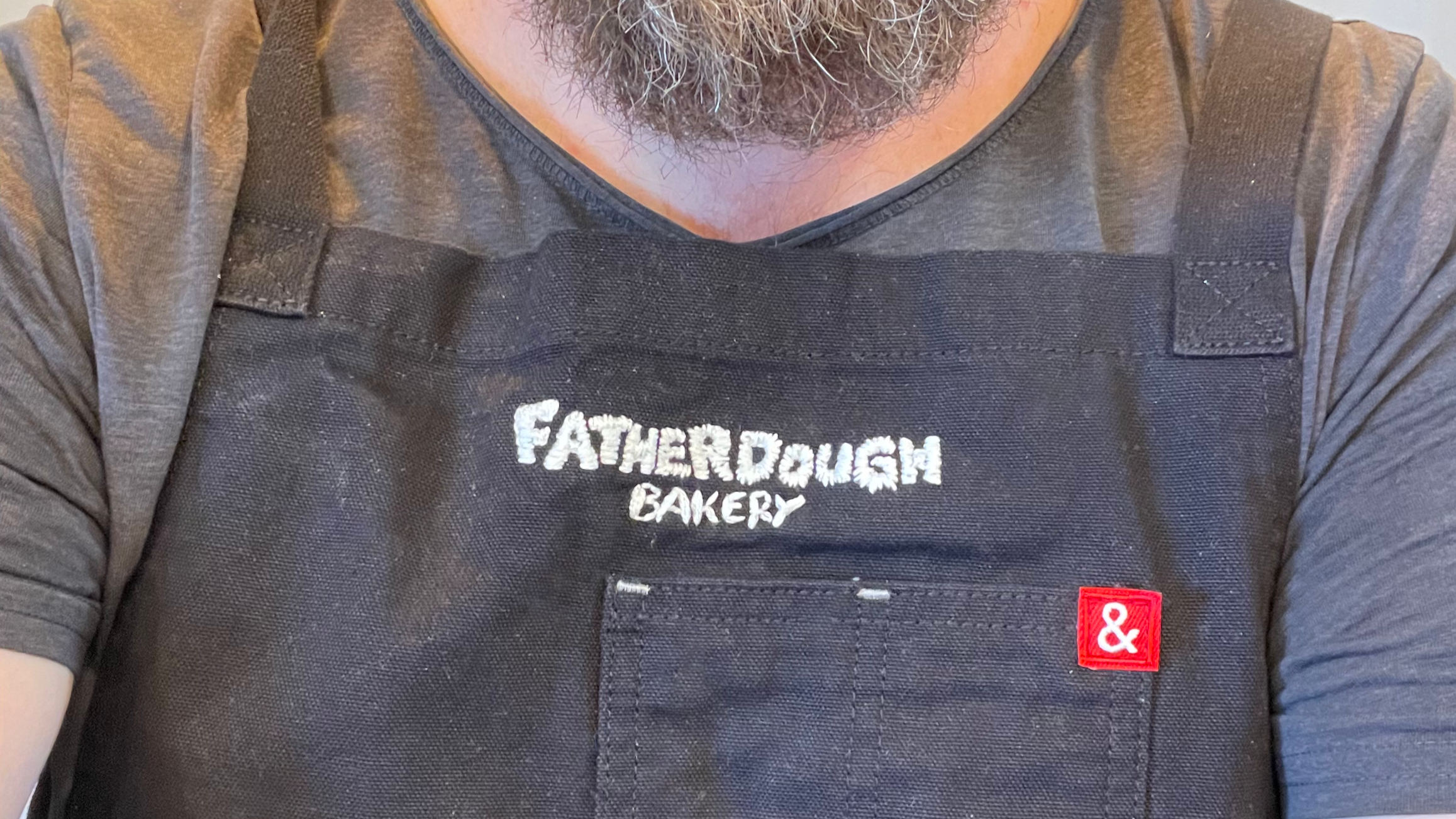 Close-up of a person wearing a black apron with the words 'Father Dough Bakery' embroidered on it, and a gray T-shirt underneath, featuring a small red and white tag.