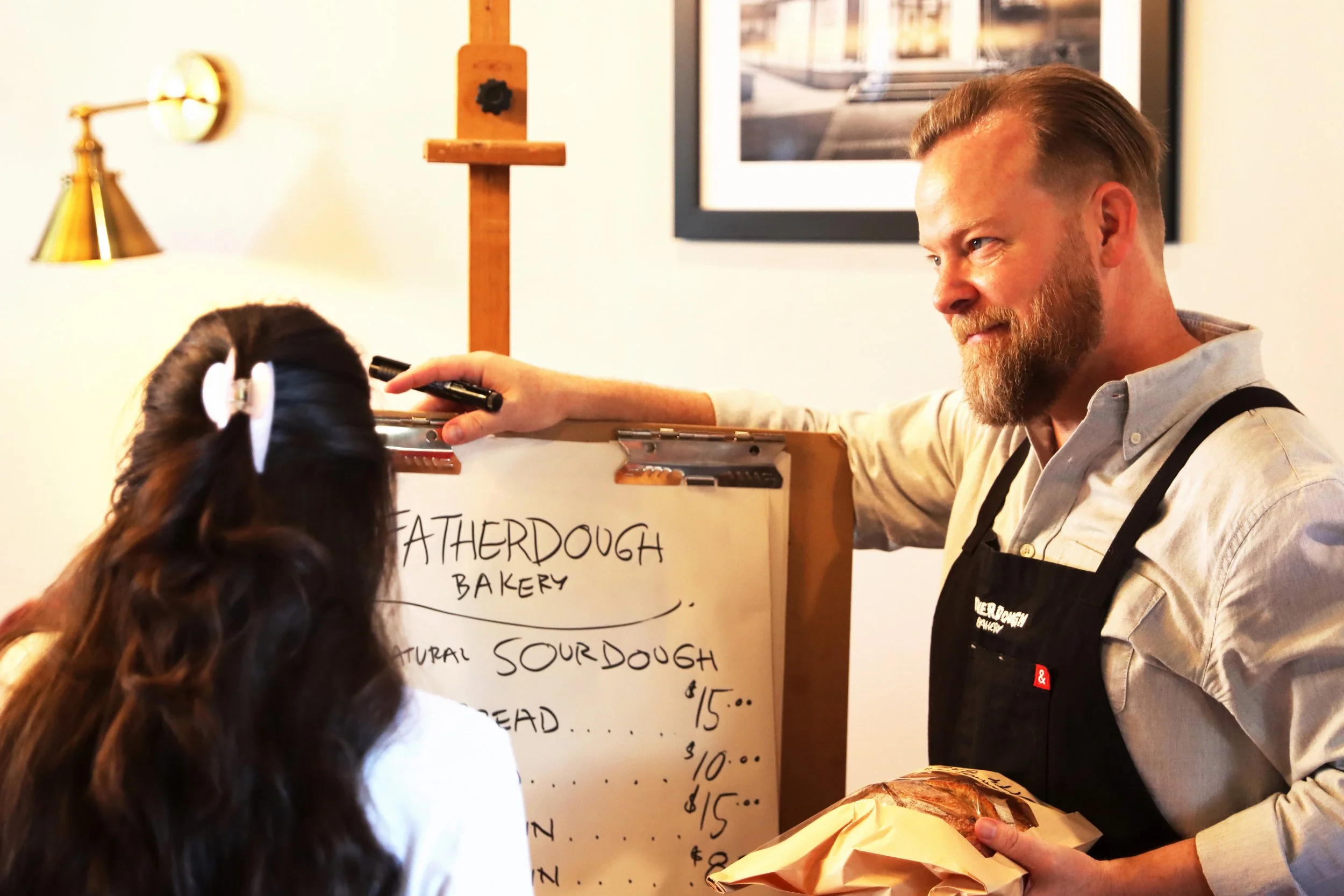 A bakery worker with a beard and apron serving a customer with long, dark hair in a ponytail at a bakery counter. The worker is smiling and holding baked goods, with a handwritten menu on a whiteboard behind them.