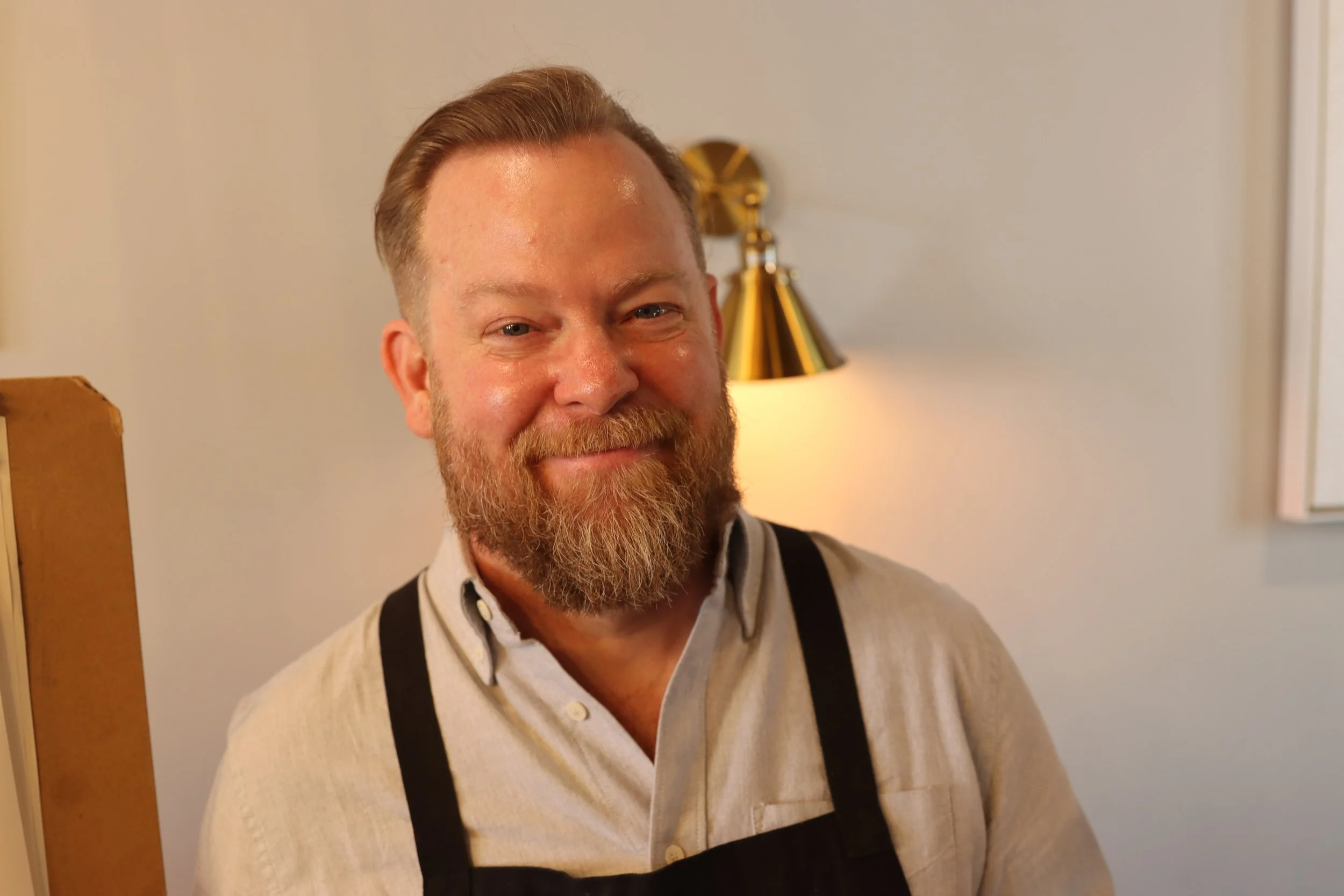 A smiling man with a beard and short hair, wearing a light-colored sweater and black suspenders, standing indoors near a wall with a lampshade in the background.