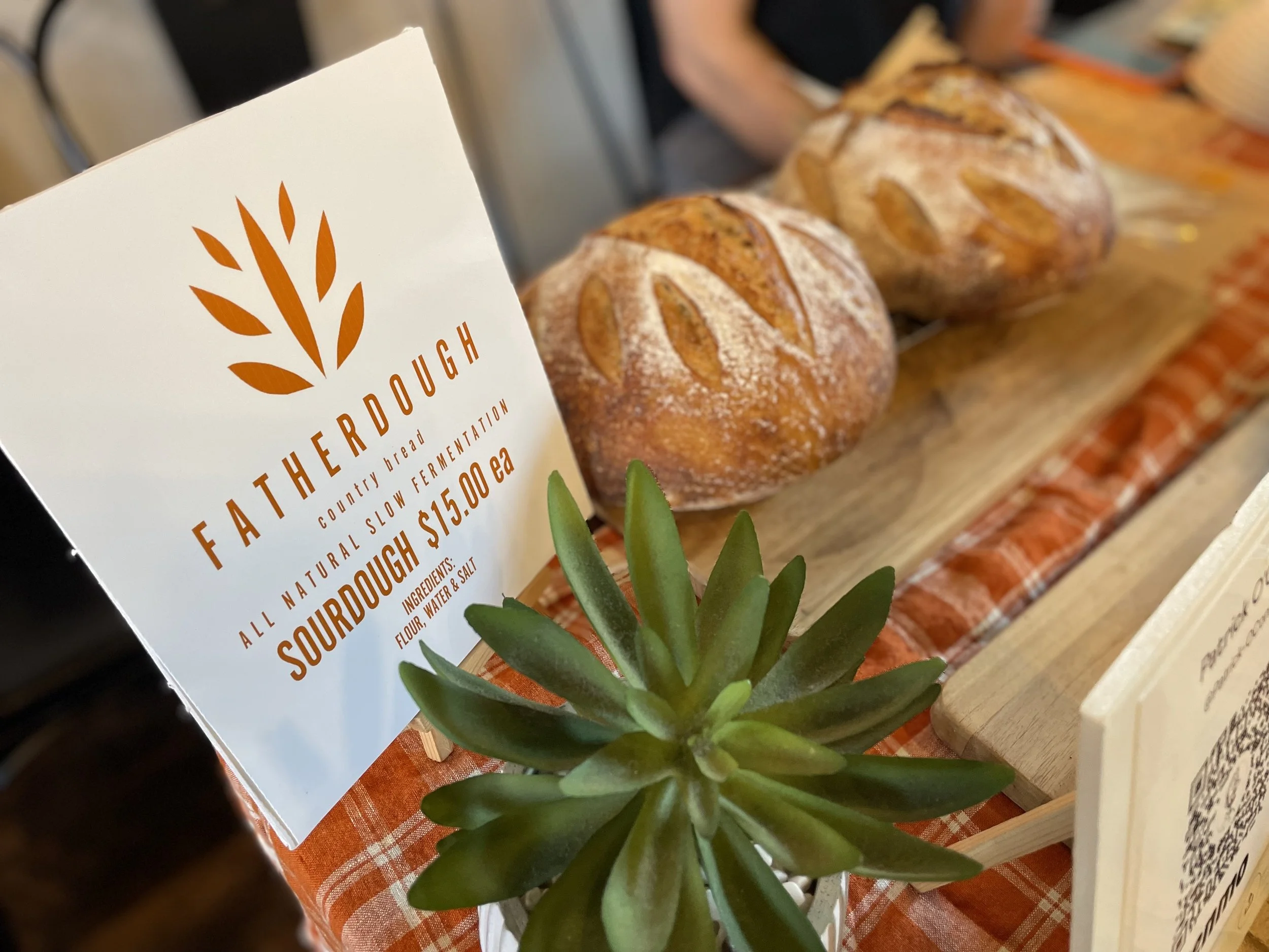 Close-up of a plant with thick green leaves placed on a table with a red gingham tablecloth. To the left, part of a white paper sign with orange text and logo that reads "FARTHEROUGH" and describes sourdough bread.