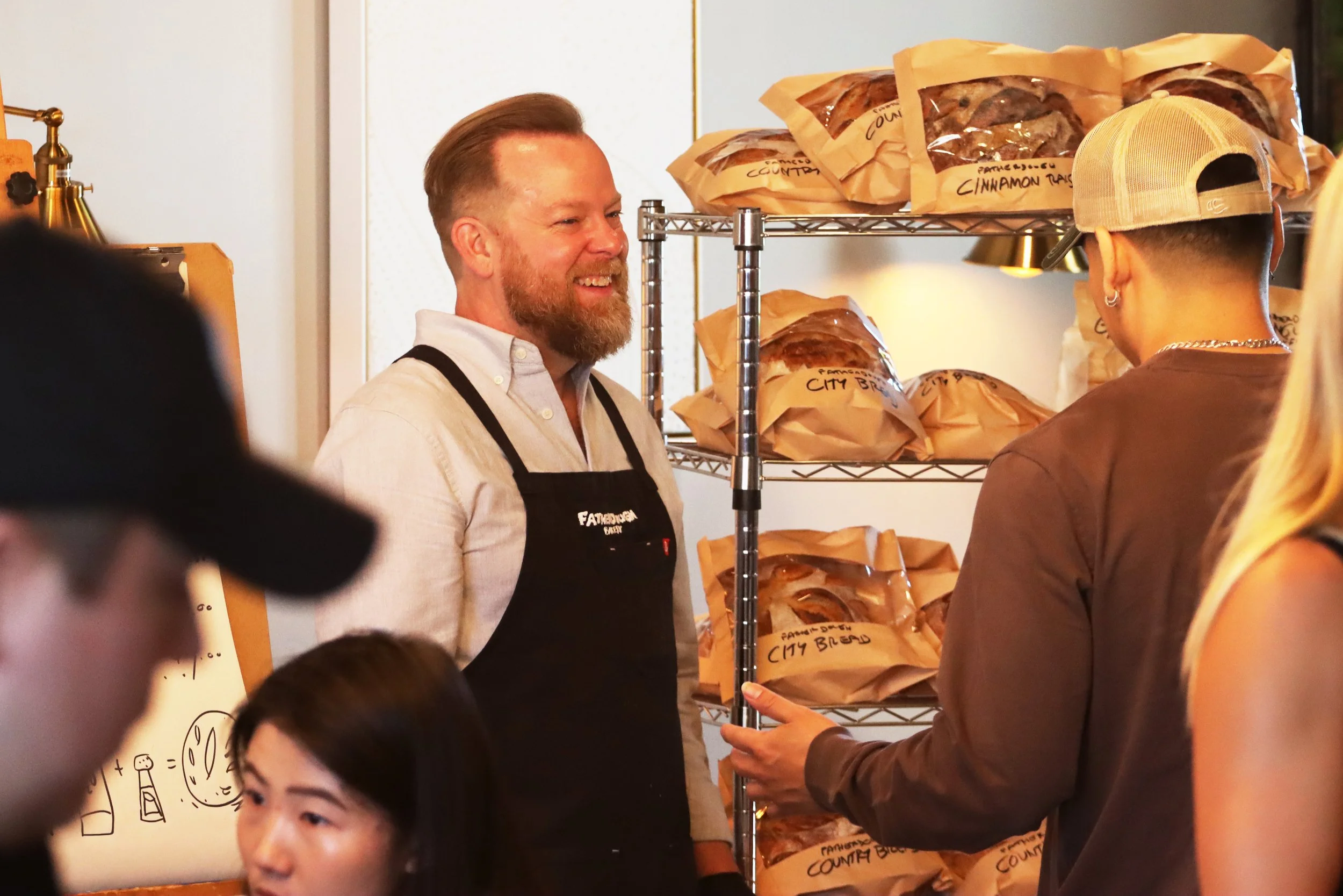 A man with a beard and a white shirt is smiling and talking to a woman wearing a beige cap and brown shirt in front of shelves with bags labeled cinnamon raisin bread. Several children are visible in the foreground.