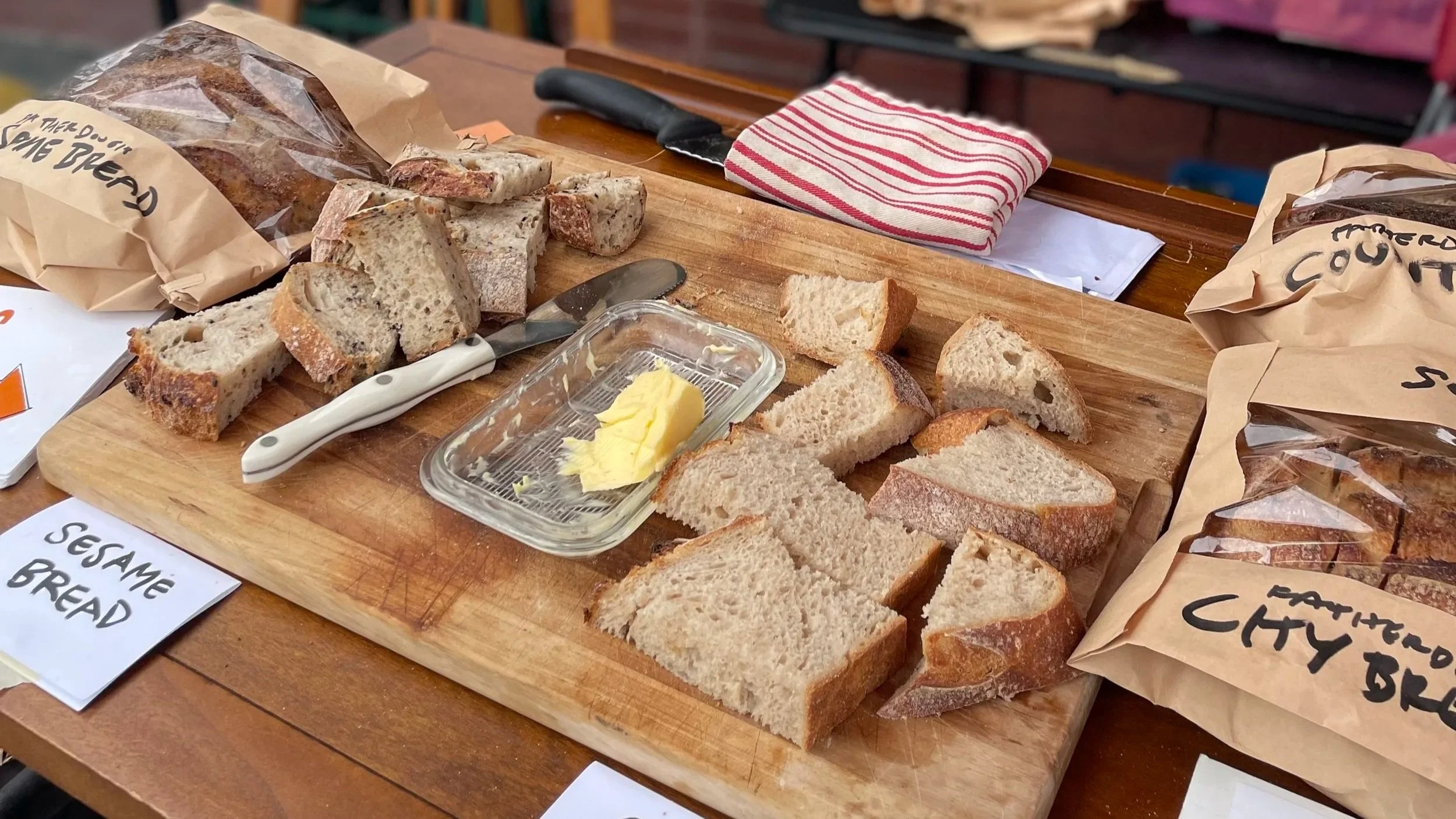 Various sliced bread on a wooden cutting board, a small dish of butter, a bread knife, and paper bags labeled with bread types on a table.