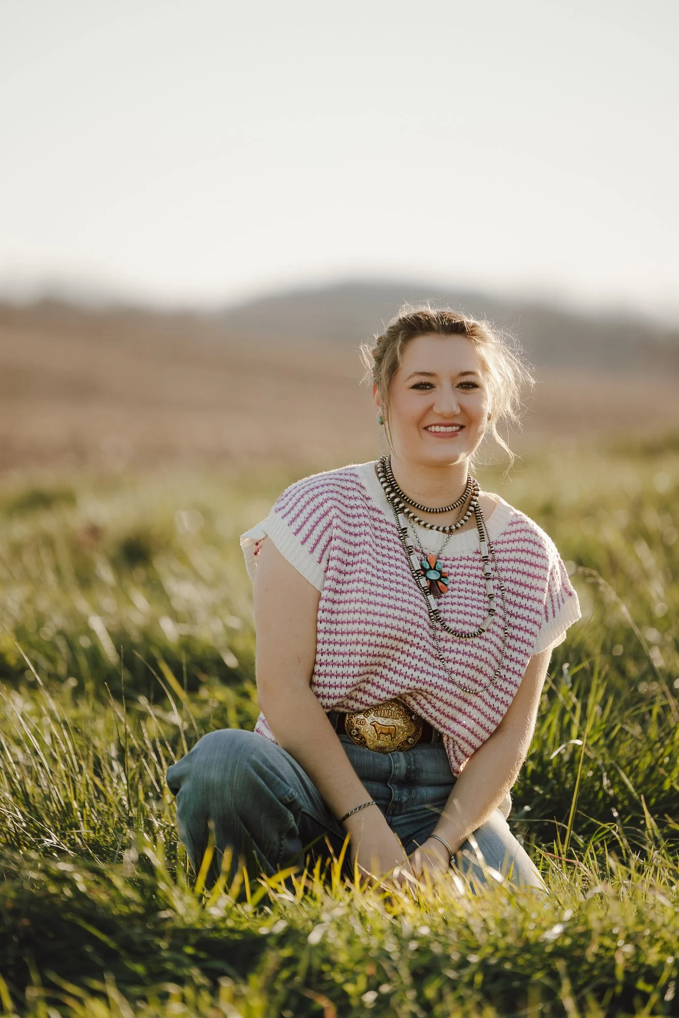 A young woman with blonde hair, smiling, sitting on grass in an outdoor field with rolling hills in the background, wearing a patterned t-shirt, jeans, and layered necklaces.