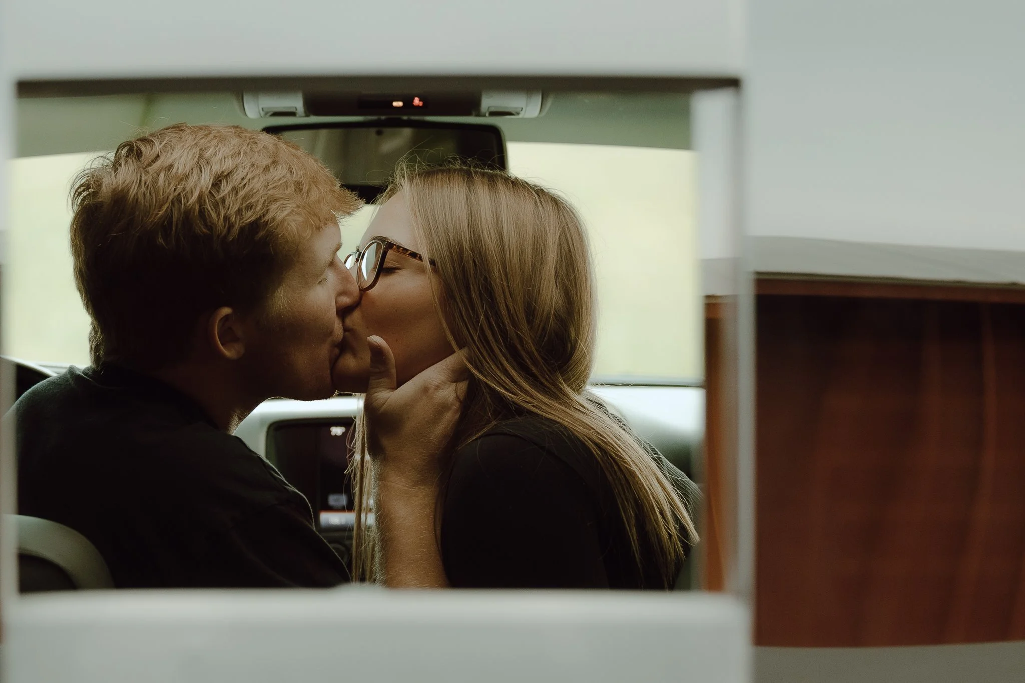 A couple sharing a kiss inside a vehicle, seen through a partially open window.