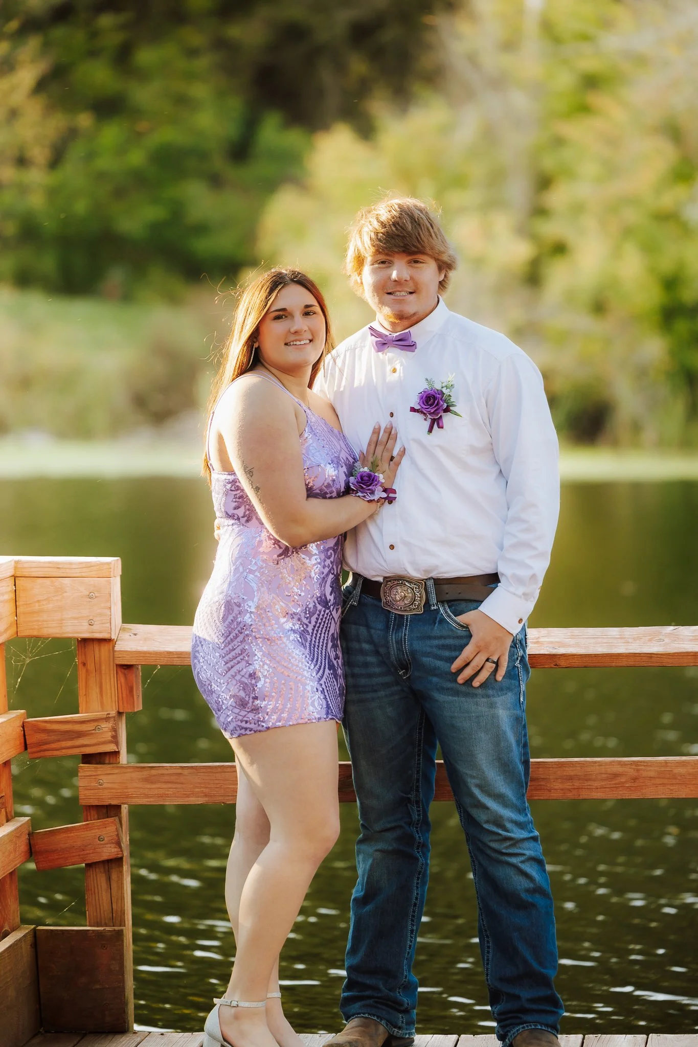 A young couple standing on a wooden dock by a lake, dressed formally with purple accents, smiling at the camera, surrounded by greenery.