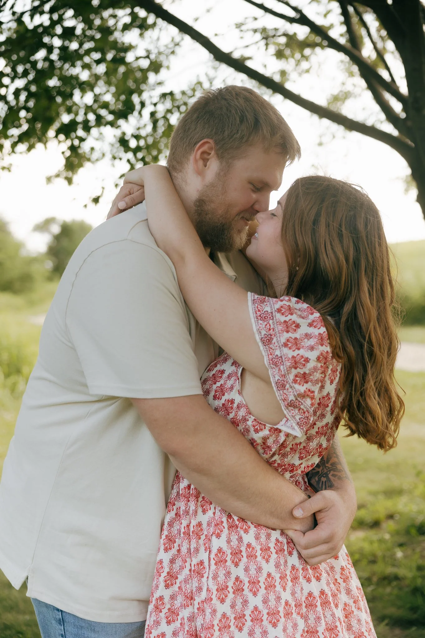 A couple hugging outdoors under a tree, smiling and touching noses.