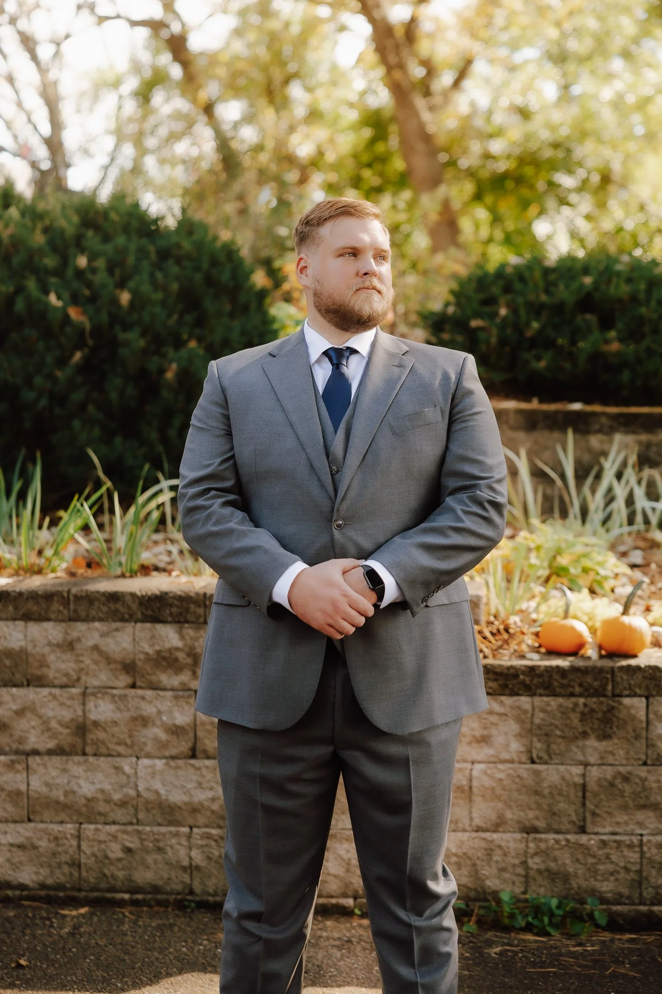 A man in a gray suit, white shirt, and navy tie stands outdoors with his hands clasped, looking to the side, with autumn trees and pumpkins in the background.
