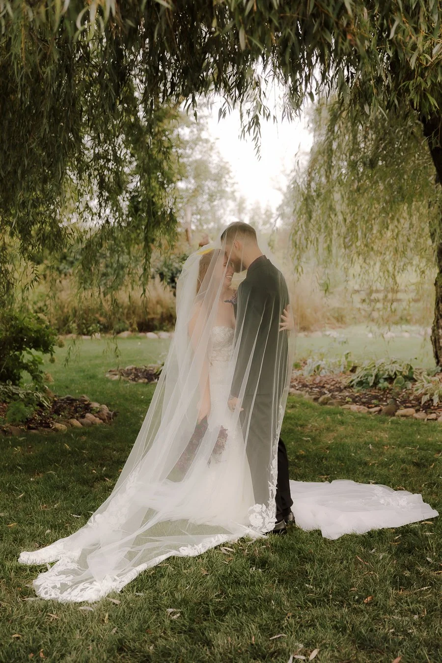 A bride and groom stand close together under a tree, smiling and touching foreheads during their wedding ceremony outdoors, with a lush green backdrop.