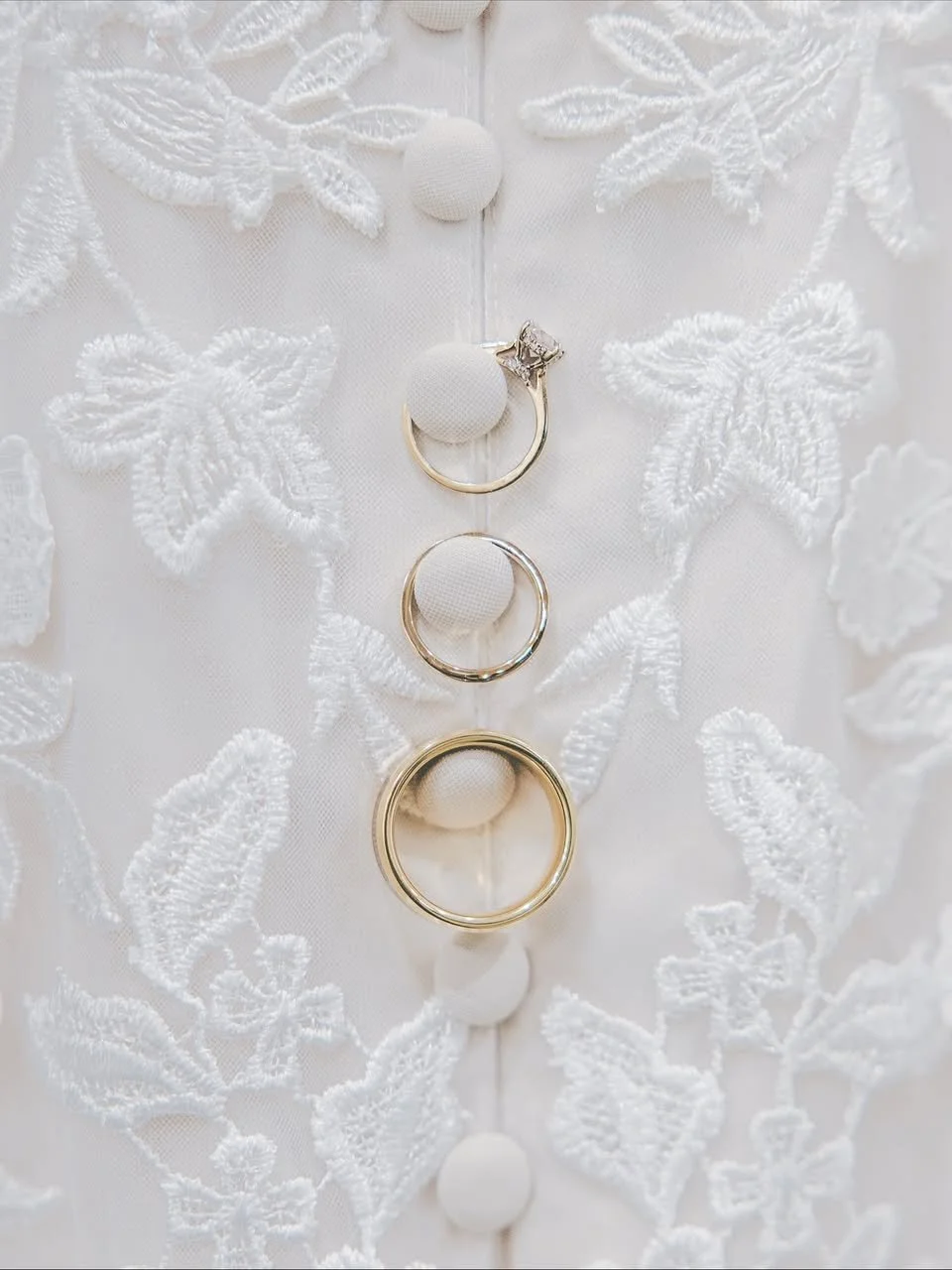 Close-up of four rings arranged vertically on a white fabric with embroidered leaf patterns and fabric-covered buttons.