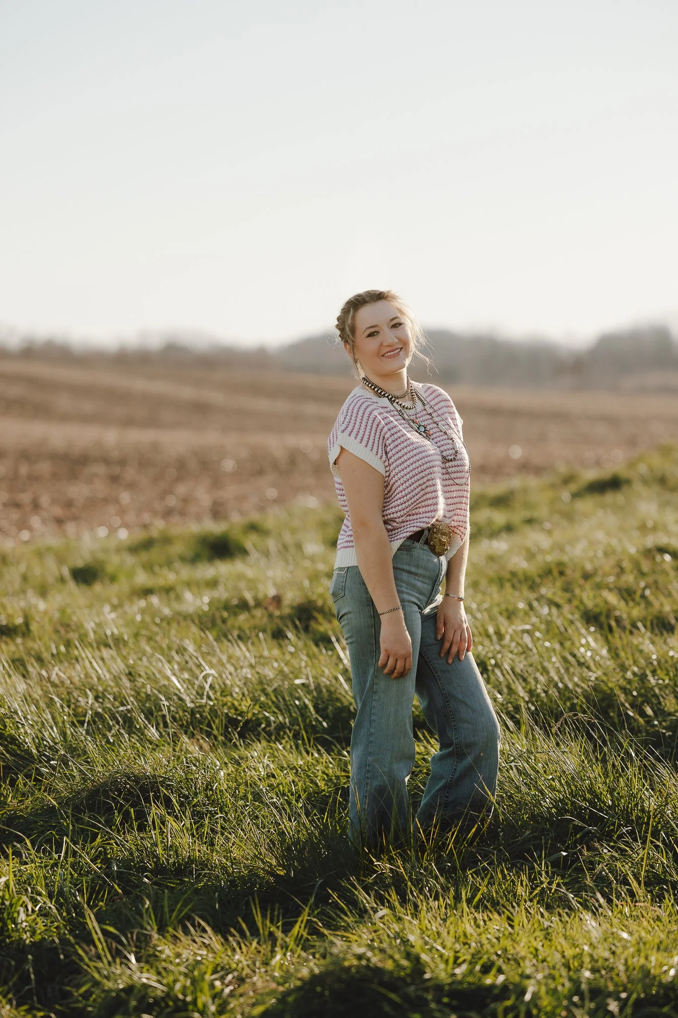 A young woman standing in a grassy field with a blurred rural landscape in the background, smiling at the camera, wearing a striped t-shirt, jeans, and layered necklaces.