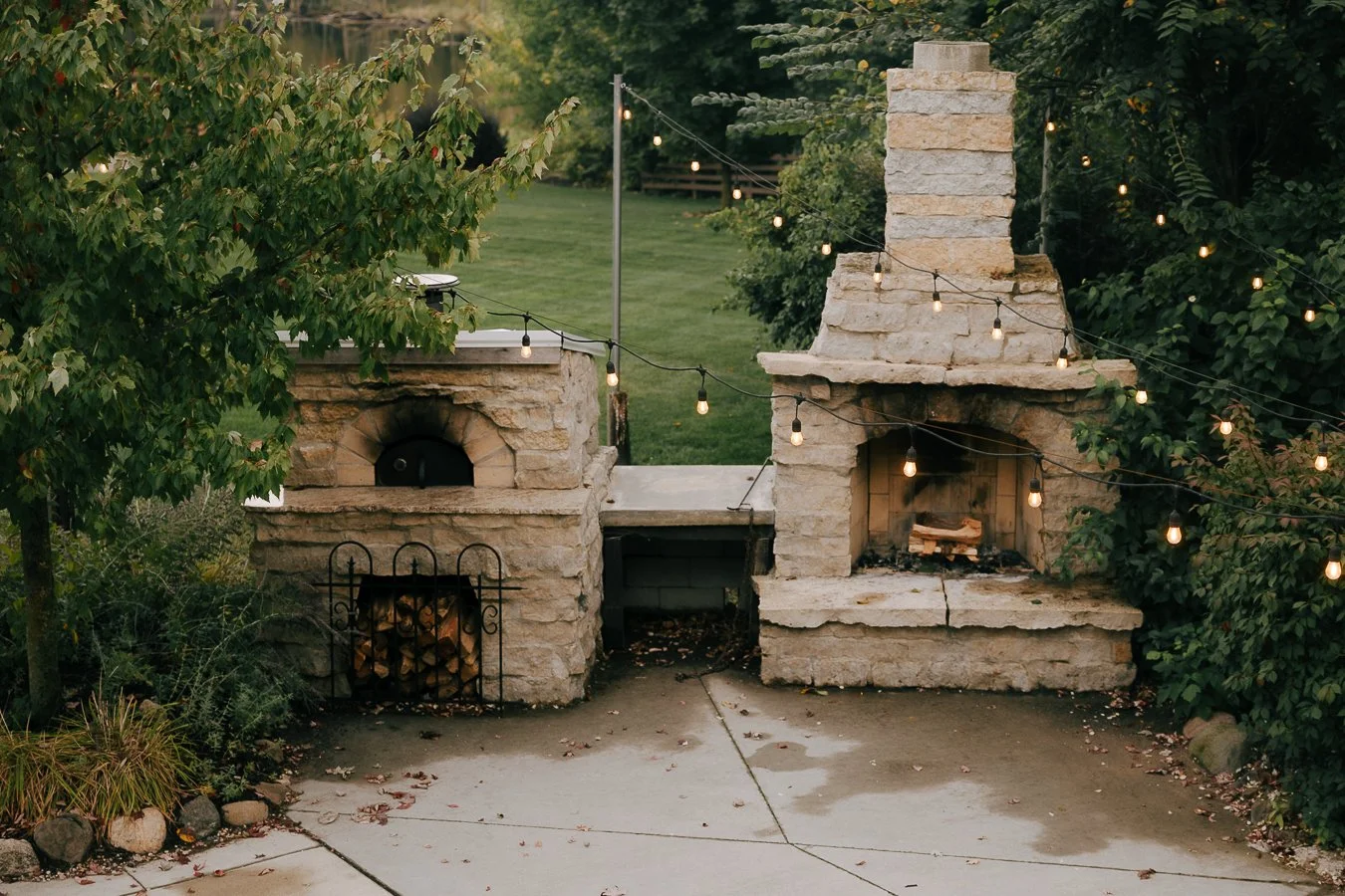 Outdoor stone fireplace with string lights, surrounded by greenery and a grassy background.