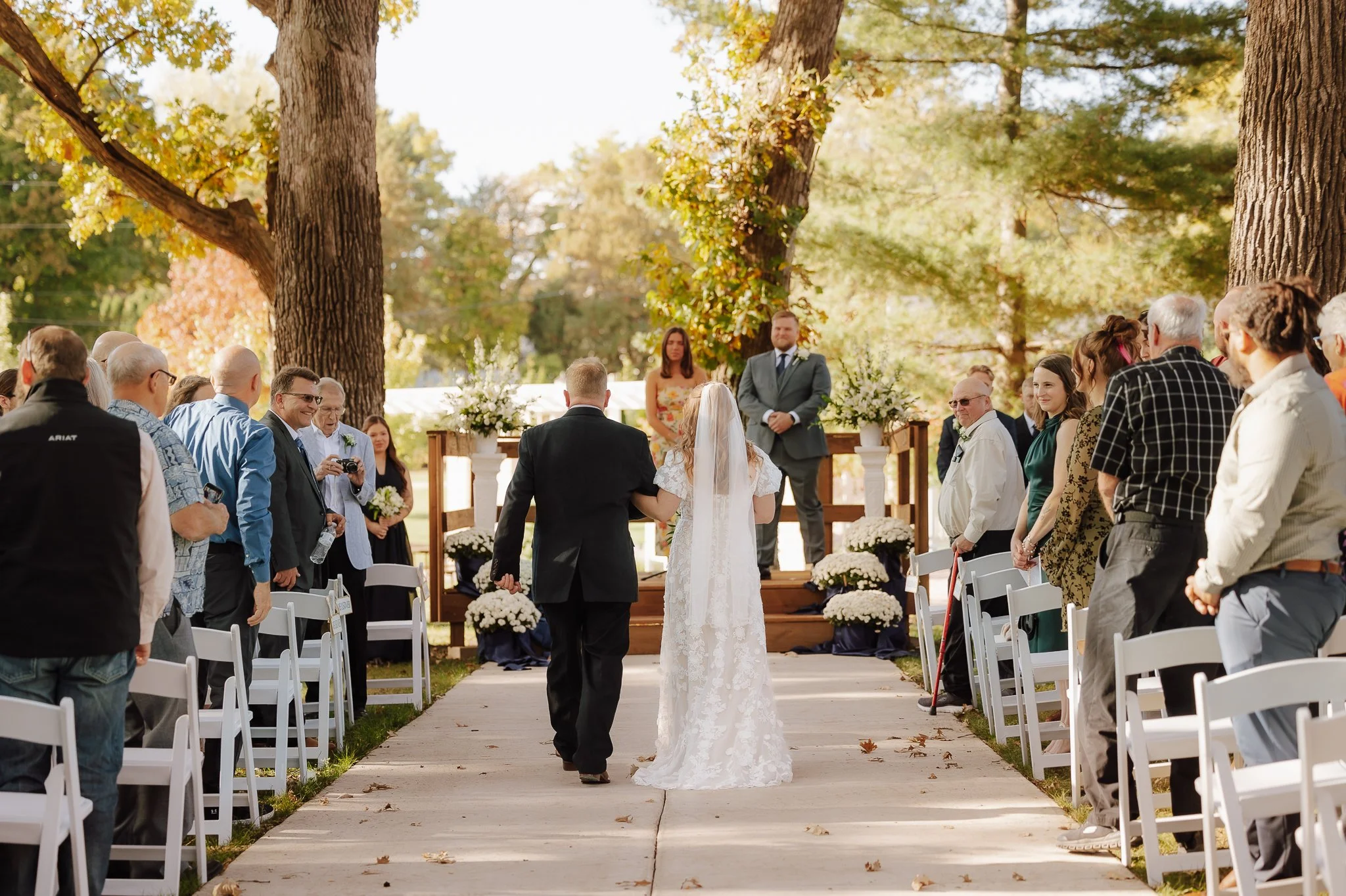 Bride and groom walking down an outdoor aisle at a wedding ceremony in a park surrounded by guests, trees, and autumn foliage.