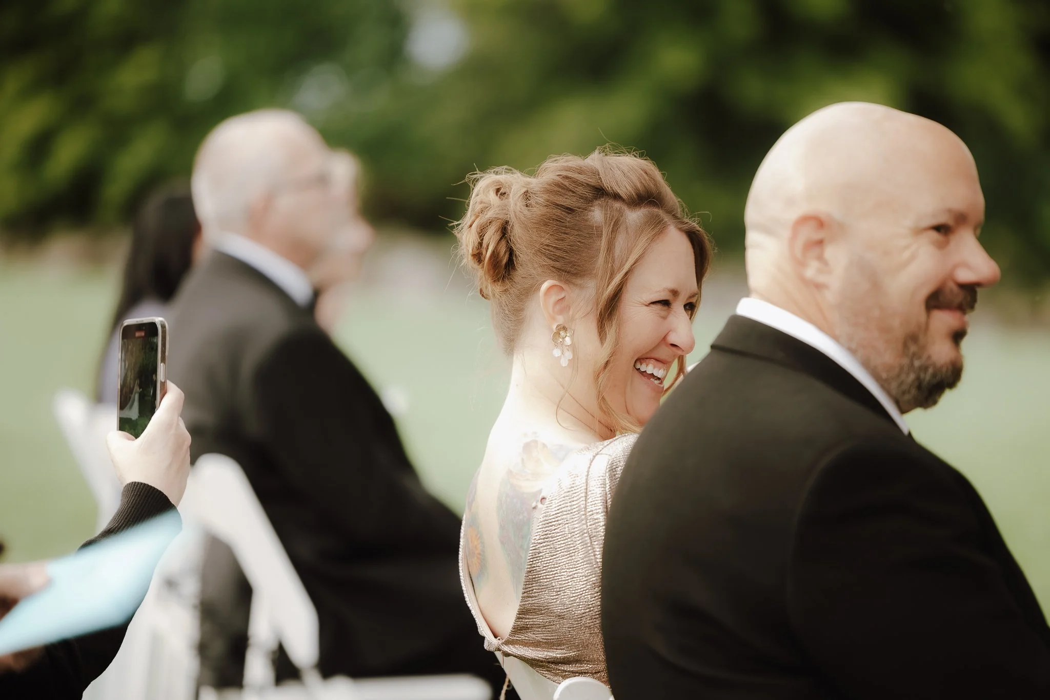 A woman with brown hair styled in an updo and earrings is smiling and laughing, seated outdoors next to a bald man in a tuxedo, with other seated guests and a green background
