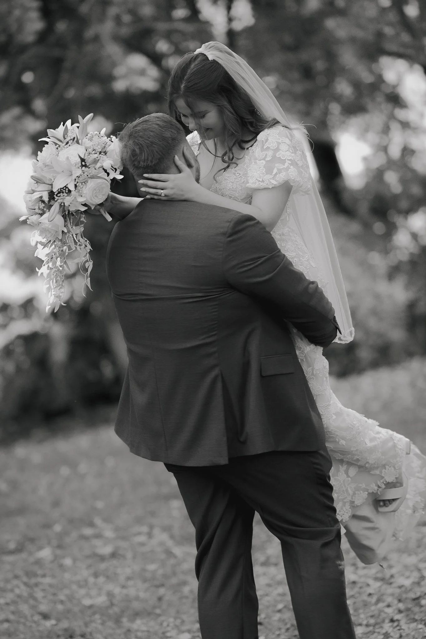 A black and white photo of a bride lifting her groom in a joyful embrace outdoors.