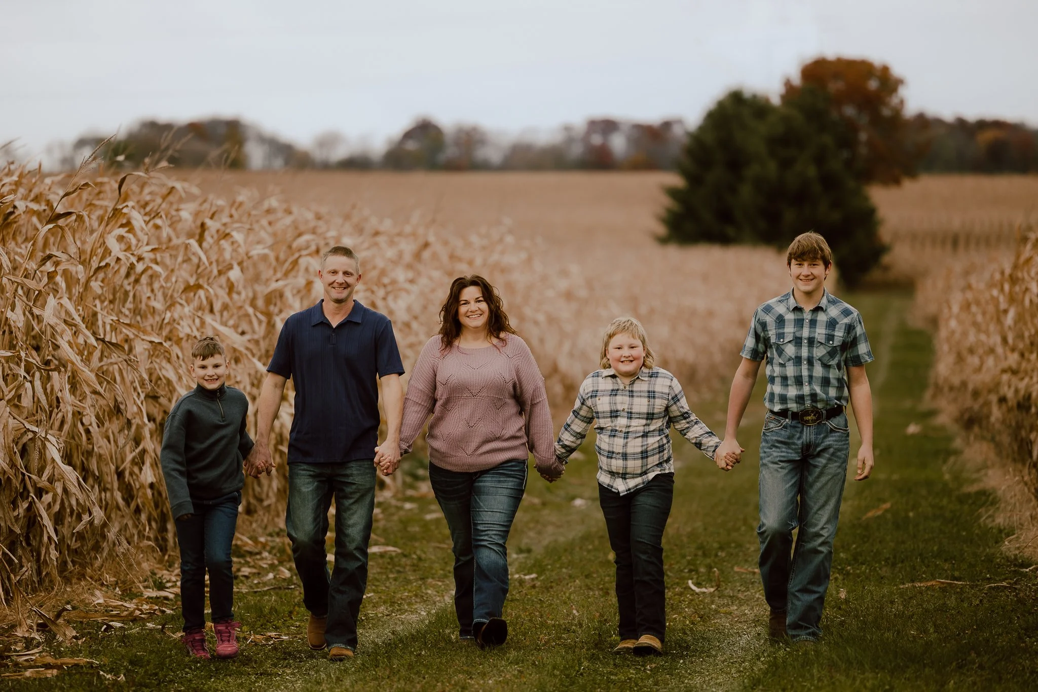 Family of five walking hand in hand through a cornfield on a fall day with colorful trees in the background.