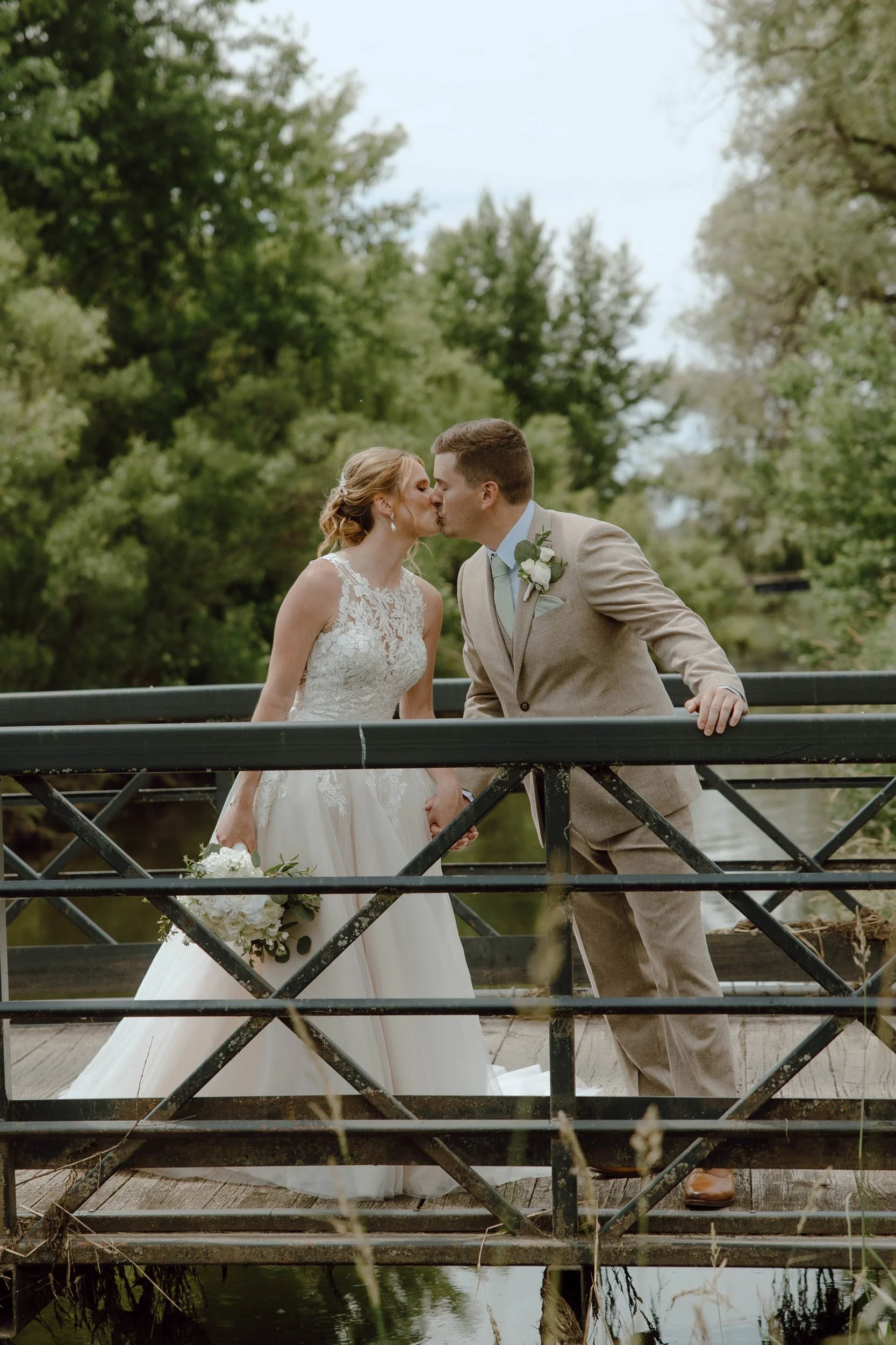 A bride and groom share a kiss on a small wooden bridge over a creek, surrounded by green trees.