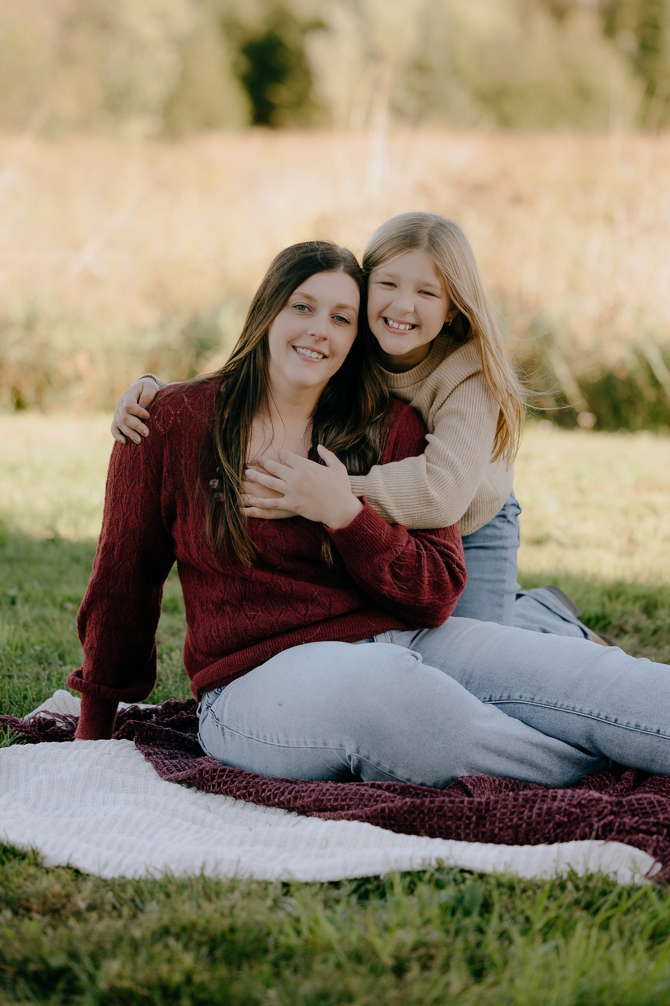 A woman and a young girl sitting on a blanket outdoors, smiling and hugging each other, with a blurred natural background.