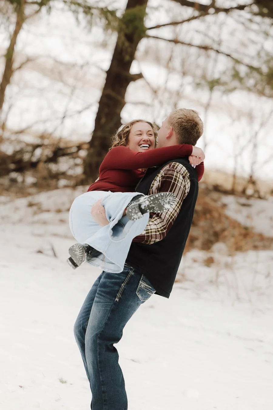 A man lifting a smiling woman in a snowy outdoor setting, with bare trees in the background.