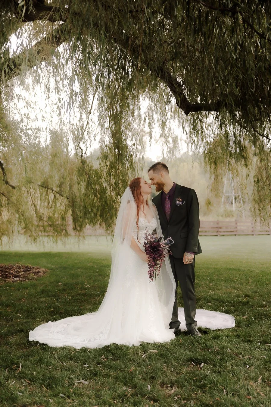 A bride and groom standing close together outdoors beneath a large tree, smiling and touching foreheads, with the bride holding a dark purple bouquet.