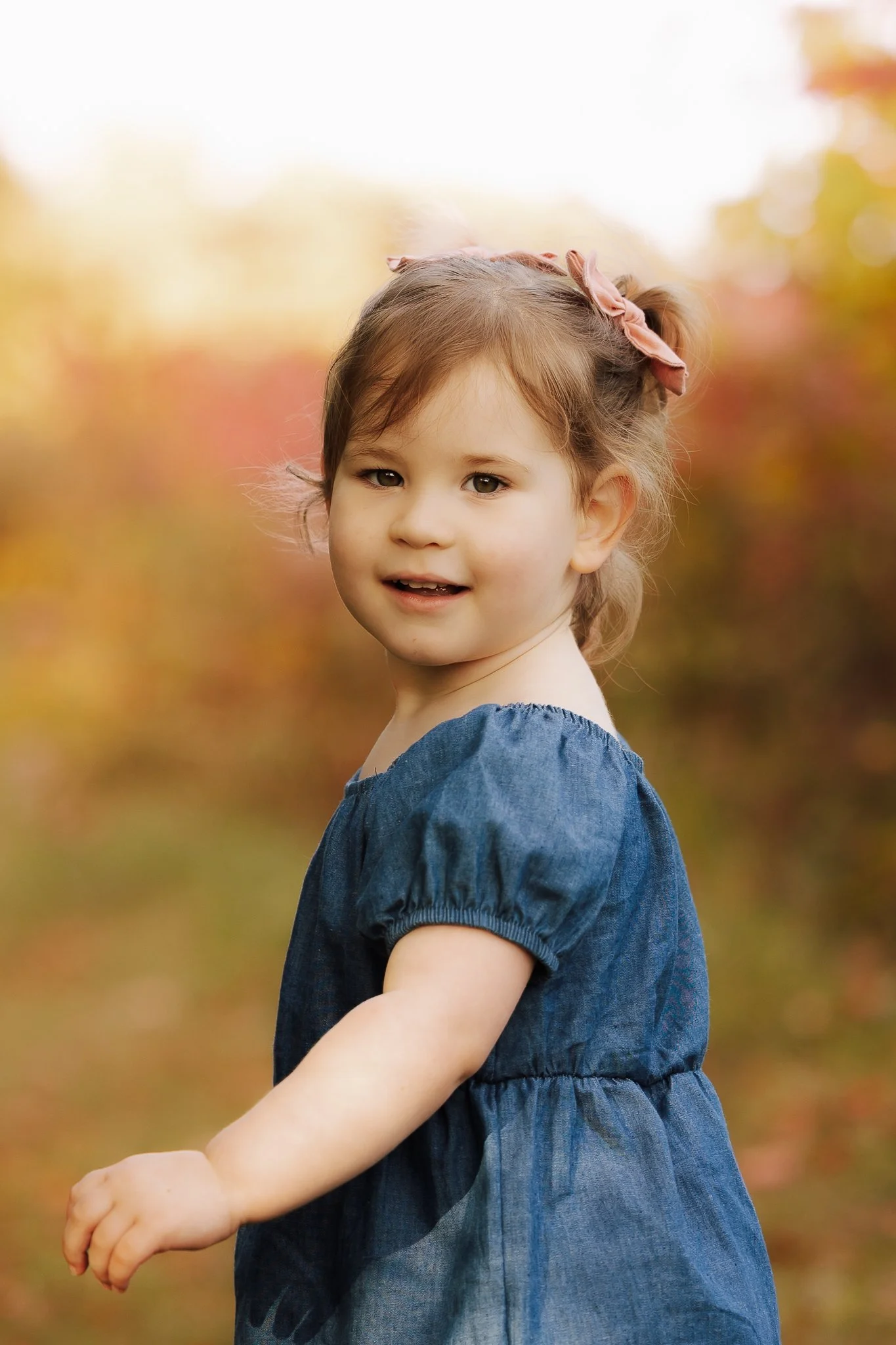 A young girl with brown hair tied with a pink bow, wearing a blue dress, standing outdoors with autumn-colored foliage background.