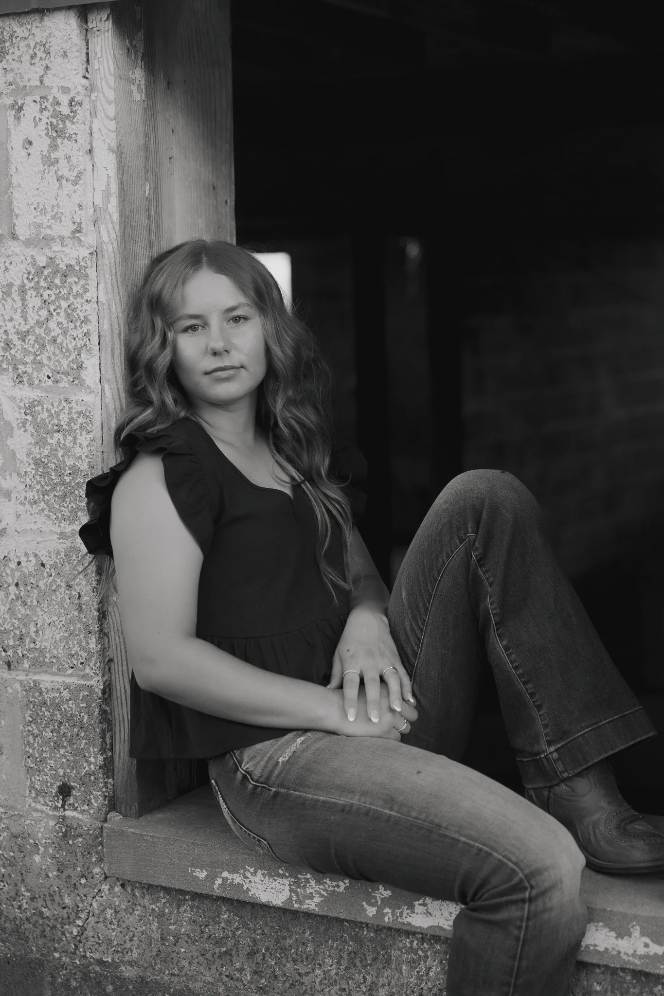 Black and white photo of a young woman with long wavy hair sitting on a windowsill, leaning against a wall with textured brick and wood.
