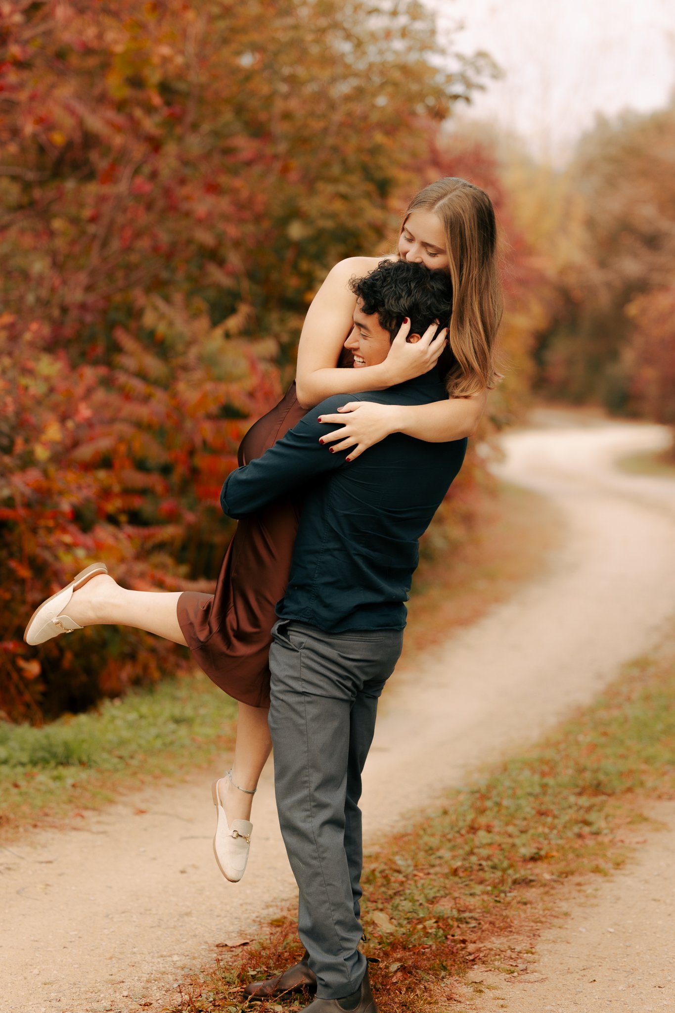 A man lifting a woman in a hug on a dirt path surrounded by autumn-colored trees.