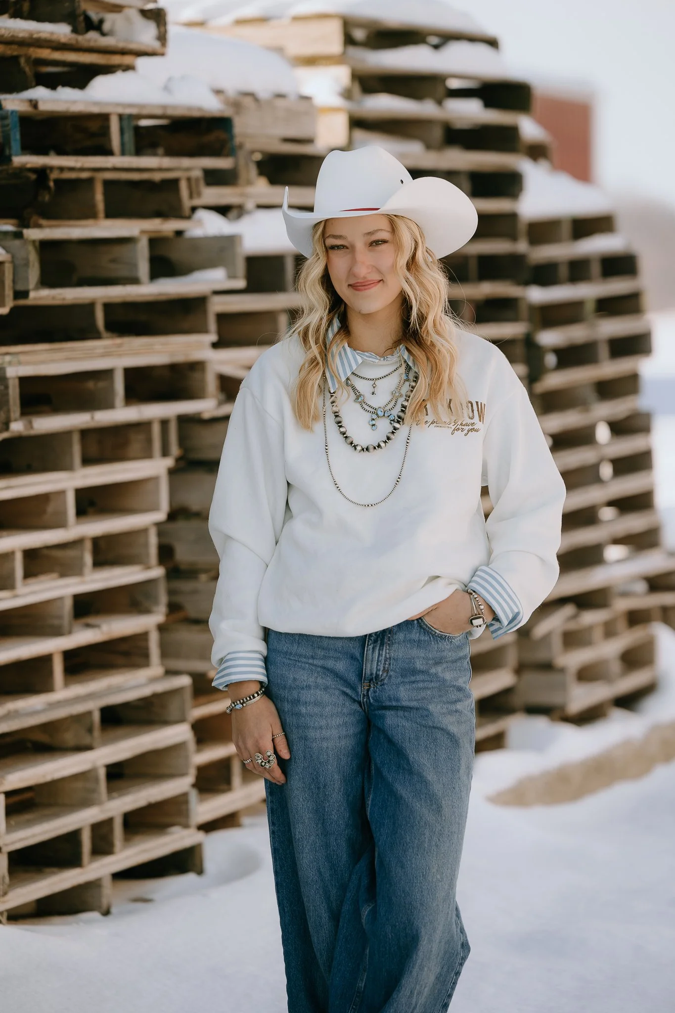 Young woman in a white cowboy hat, wearing layered necklaces, a white sweatshirt, and blue jeans, standing outdoors near stacked wooden pallets in snow.