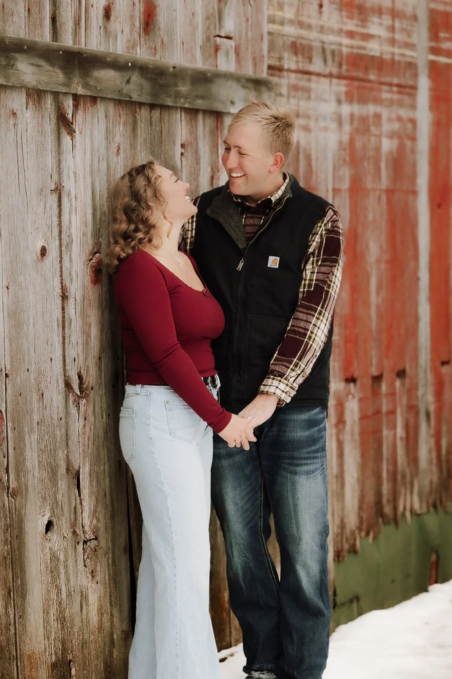 A couple standing close, holding hands, smiling at each other near a weathered wooden barn, with snow on the ground.