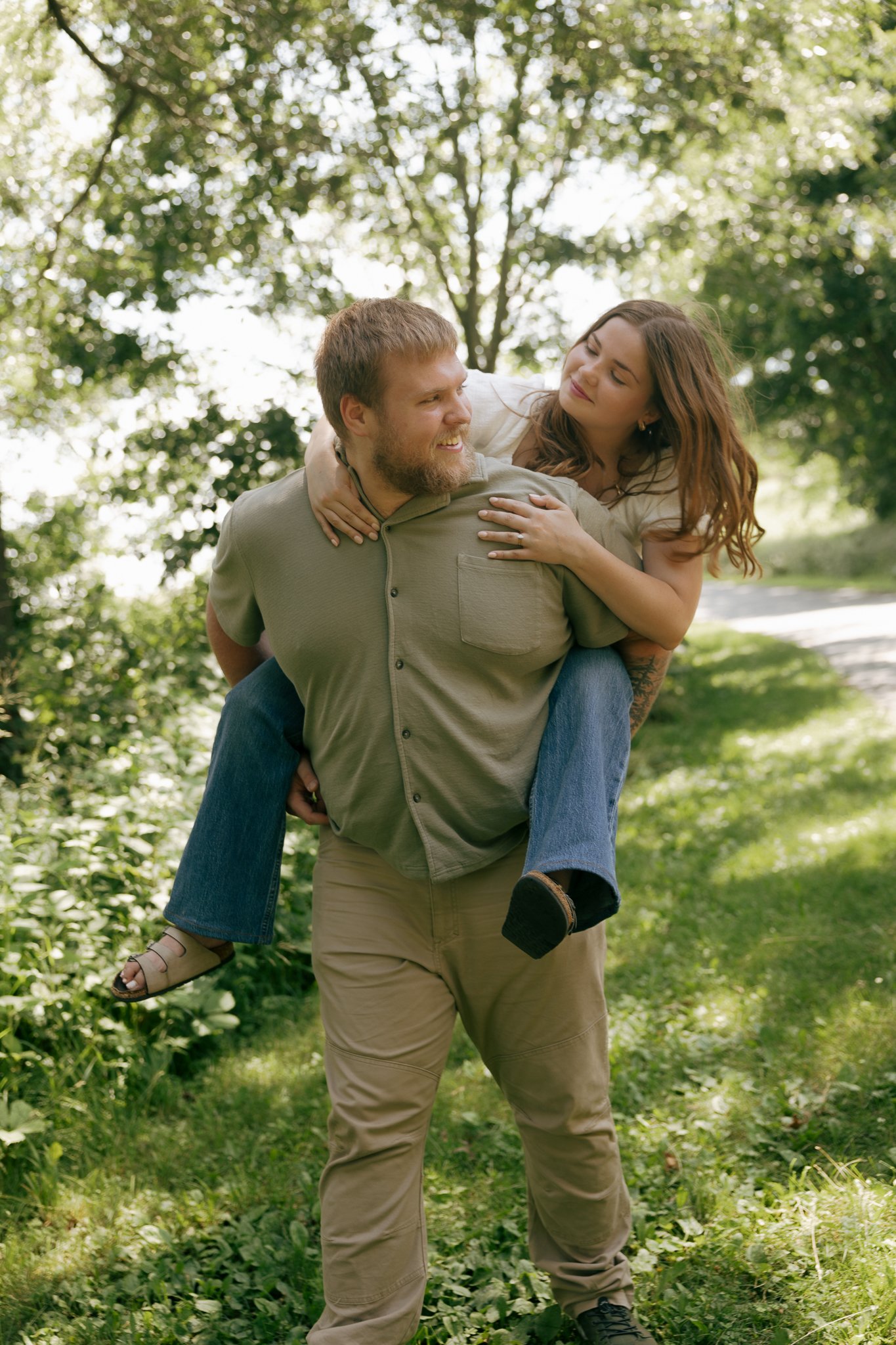 A man giving a woman a piggyback ride through a lush, green park with sunlight filtering through the trees.