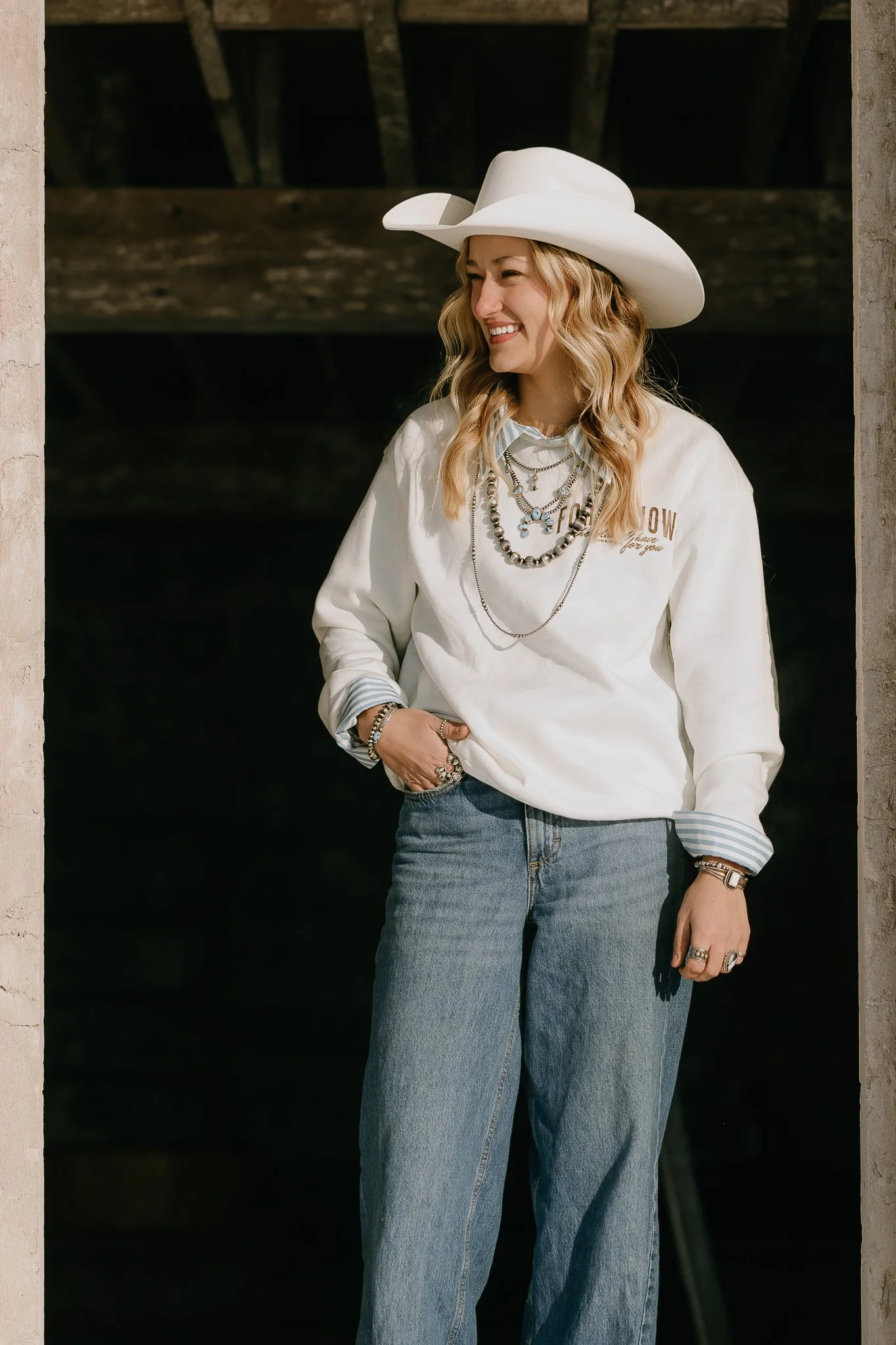 A woman standing outdoors near a structure, wearing a white cowboy hat, a white sweatshirt with embroidery, layered jewelry, and blue jeans, smiling and looking to her right.