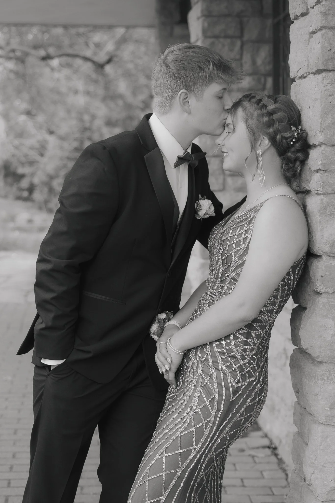 A black and white photo of a young couple in formal attire, with the man kissing the woman's forehead. They are standing outdoors next to a brick wall, and the woman is wearing a beaded gown and jewelry, smiling with her eyes closed.