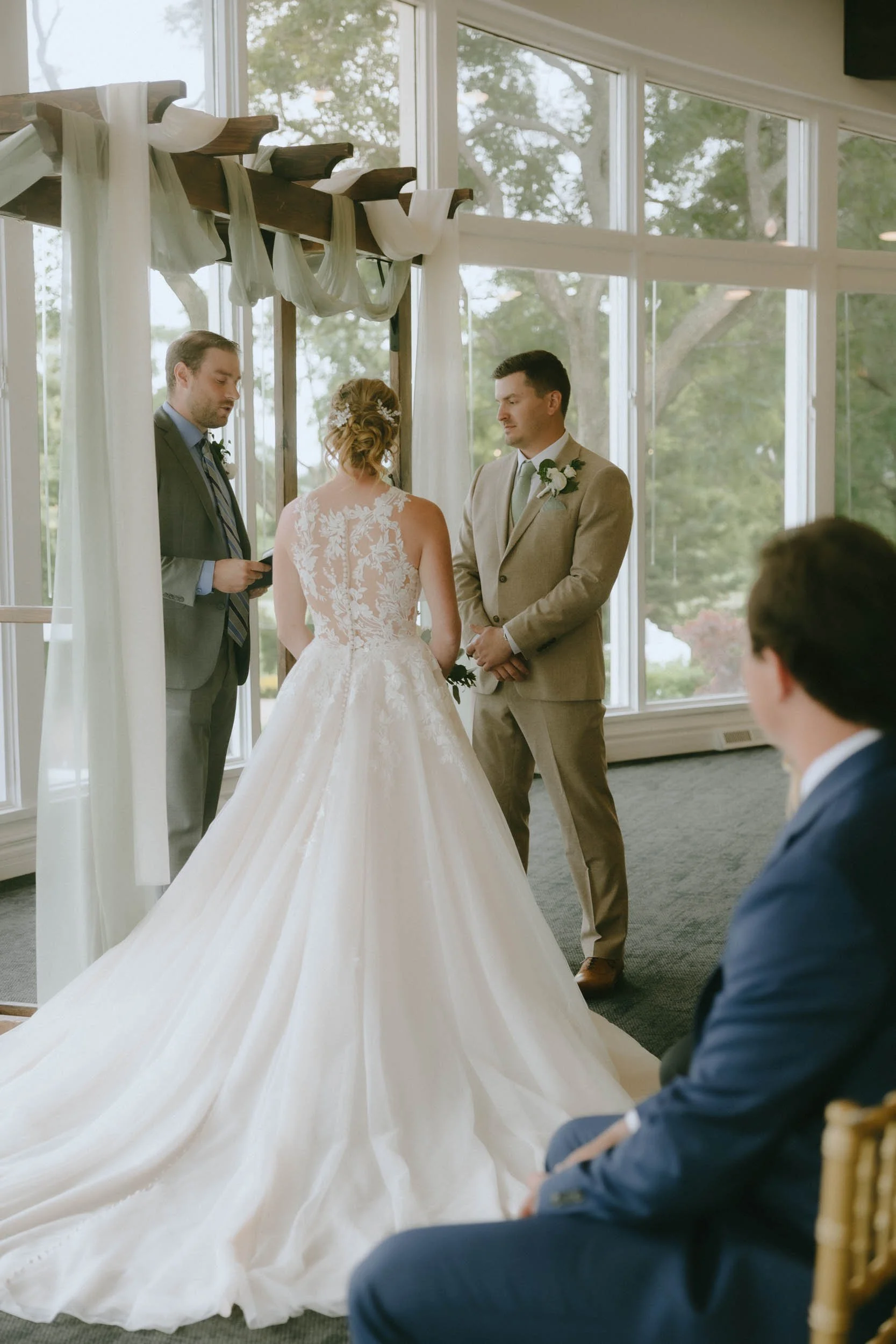 Bride and groom exchanging vows during a wedding ceremony indoors, with officiant and a seated guest in the background.