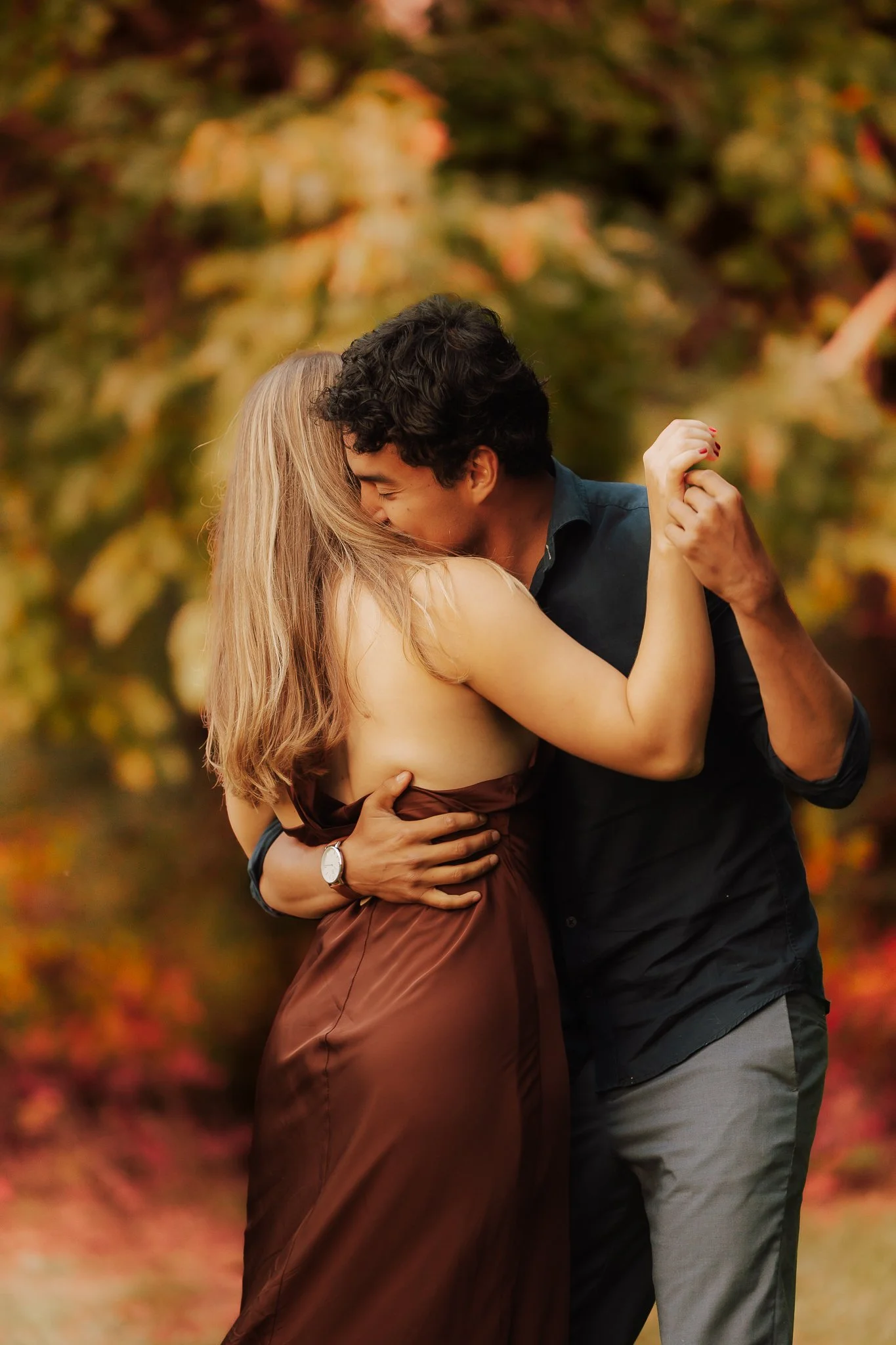 A couple dancing closely outdoors during autumn, with trees and colorful leaves in the background, embracing each other affectionately.