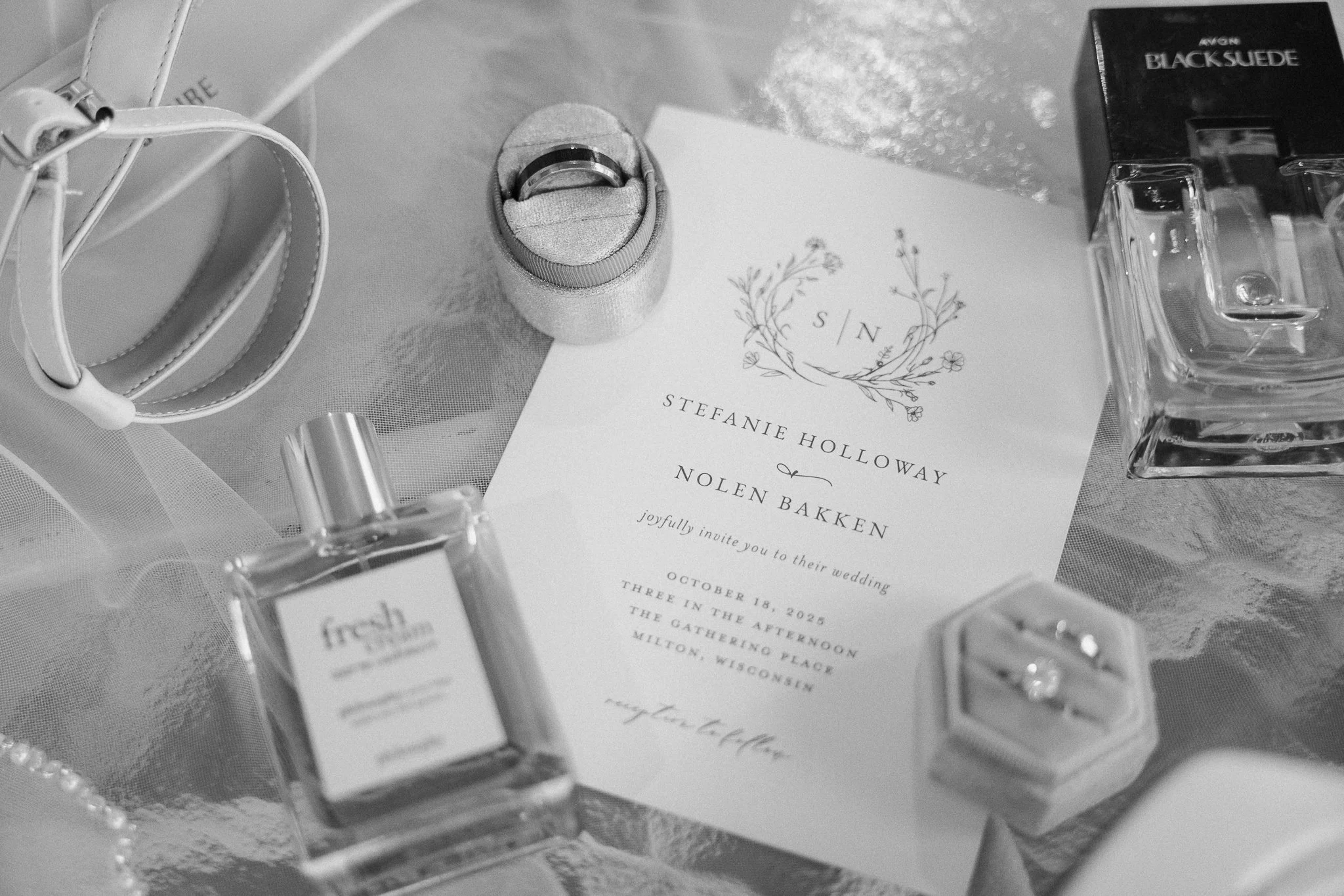 A wedding invitation surrounded by perfume bottles, rings in a box, and jewelry on a table.