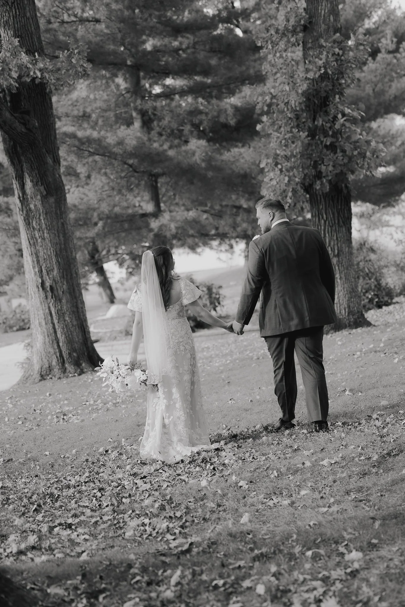 A black and white photograph of a bride and groom holding hands, walking through a park with tall trees, leaves on the ground, and a serene outdoor setting.