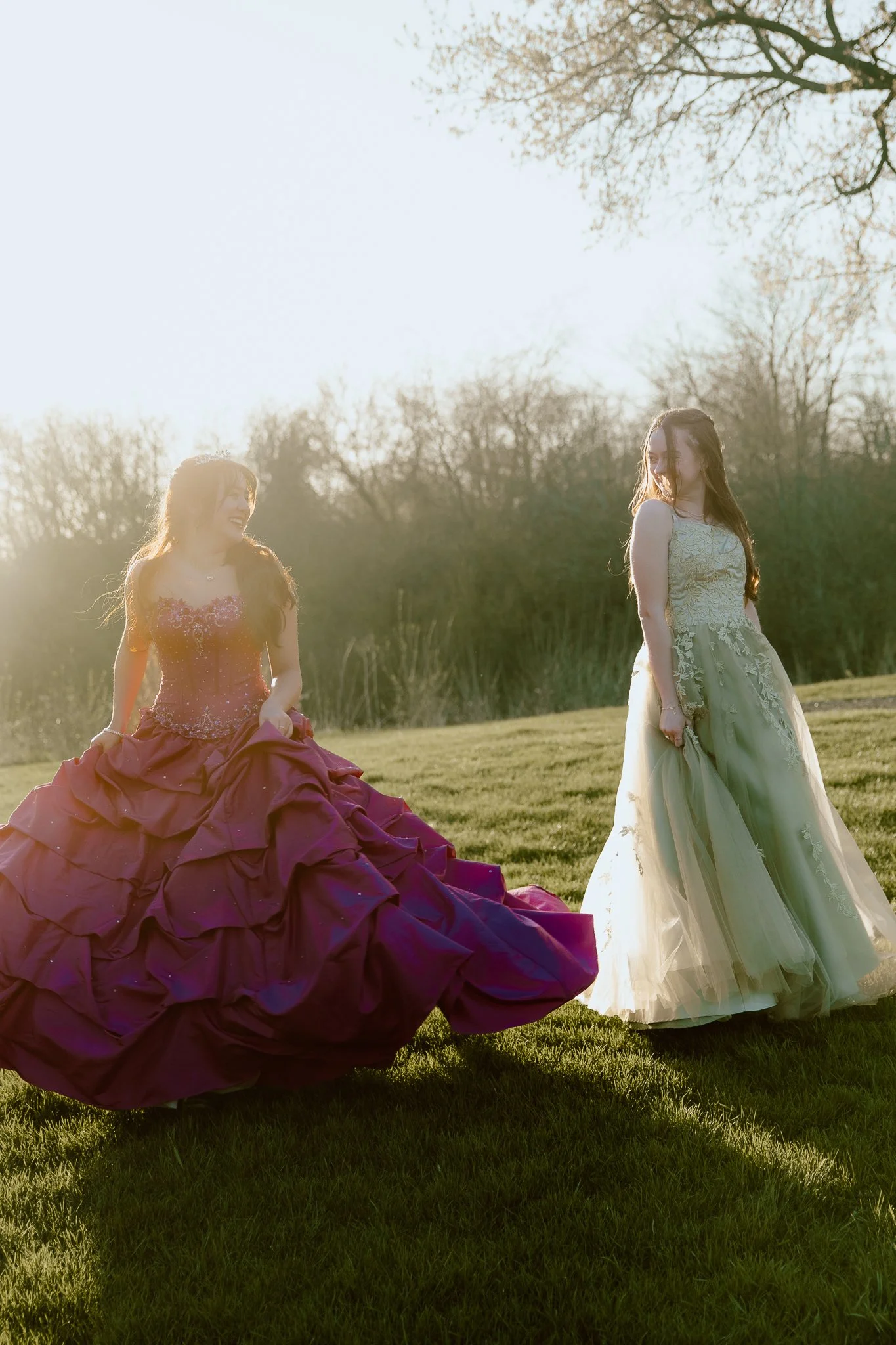 Two women in formal gowns, one in a maroon dress and the other in a pale green dress, standing on a grassy field with trees in the background, during sunset.