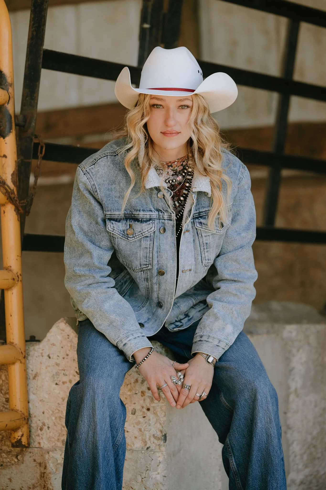 A woman wearing a white cowboy hat, denim jacket, and jewelry, sitting on a stone block in front of a metal staircase.