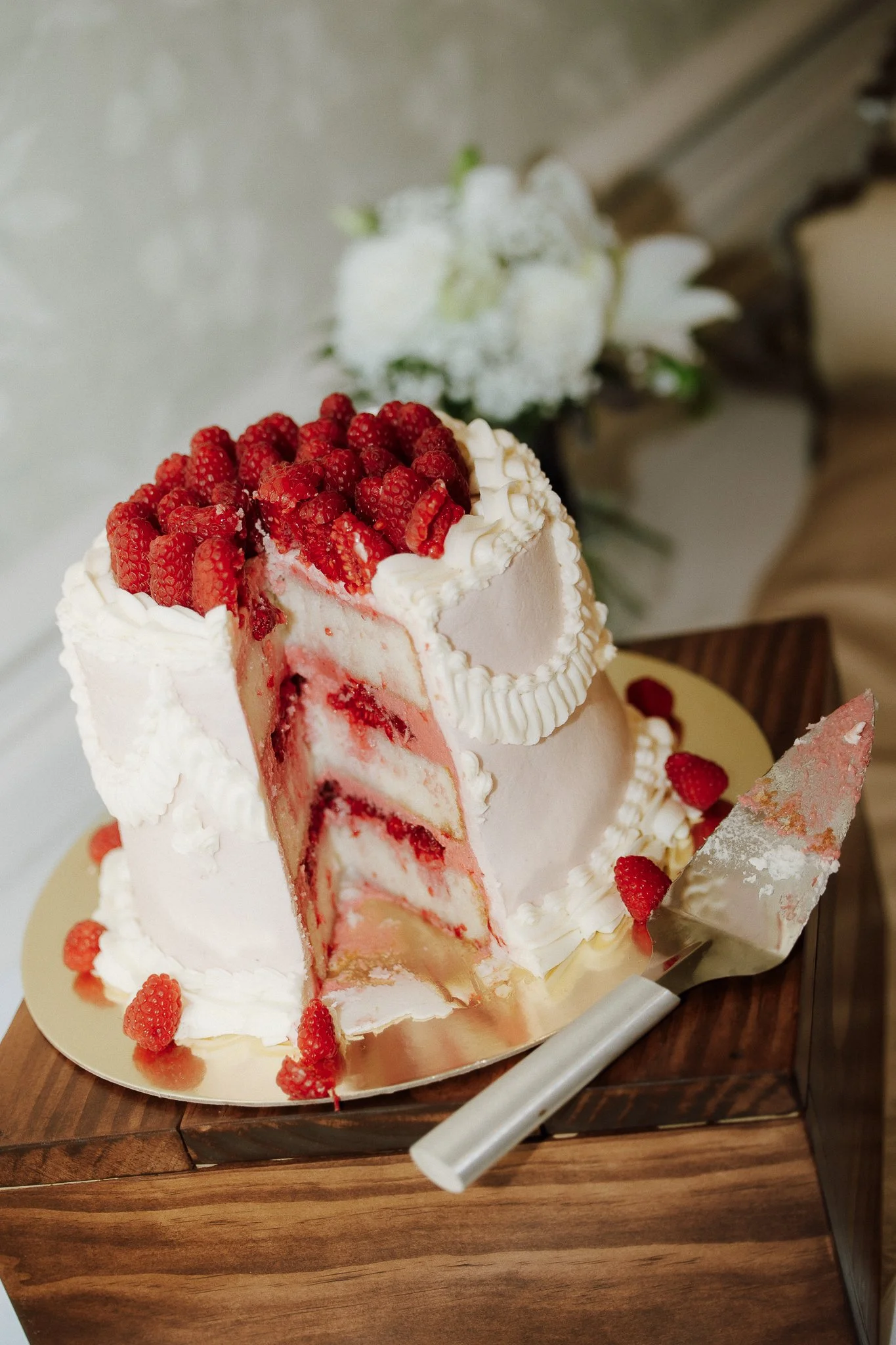A layered strawberry and cream cake with sliced strawberries on top, partially sliced to reveal layers of cream and strawberries inside, sitting on a golden cake board on a wooden surface.