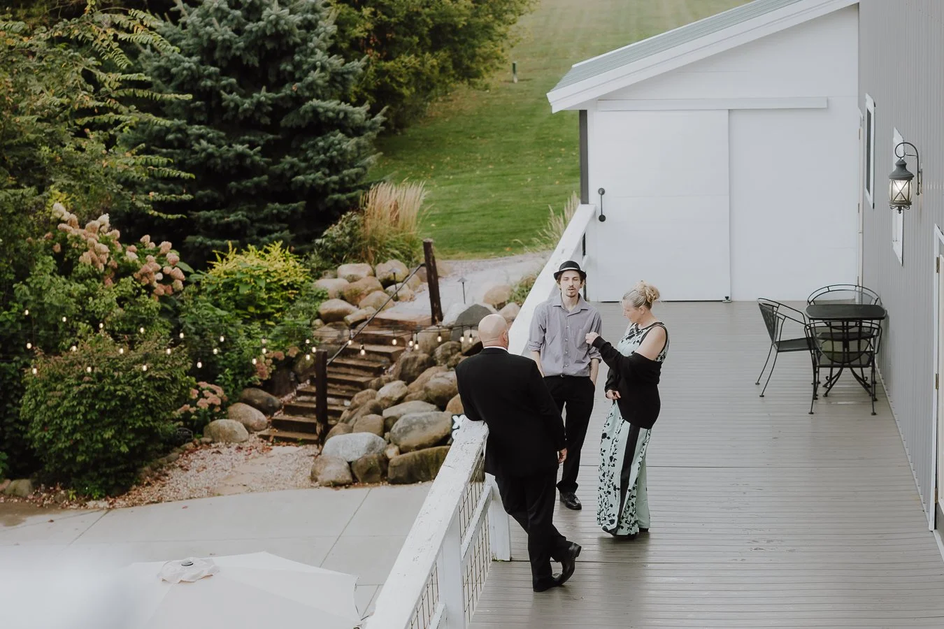 Three people stand on a wooden deck having a conversation, with one man in a suit and two others dressed casually. The deck is attached to a white house with outdoor furniture and a lantern. There are stairs leading down to a garden with rock landsca