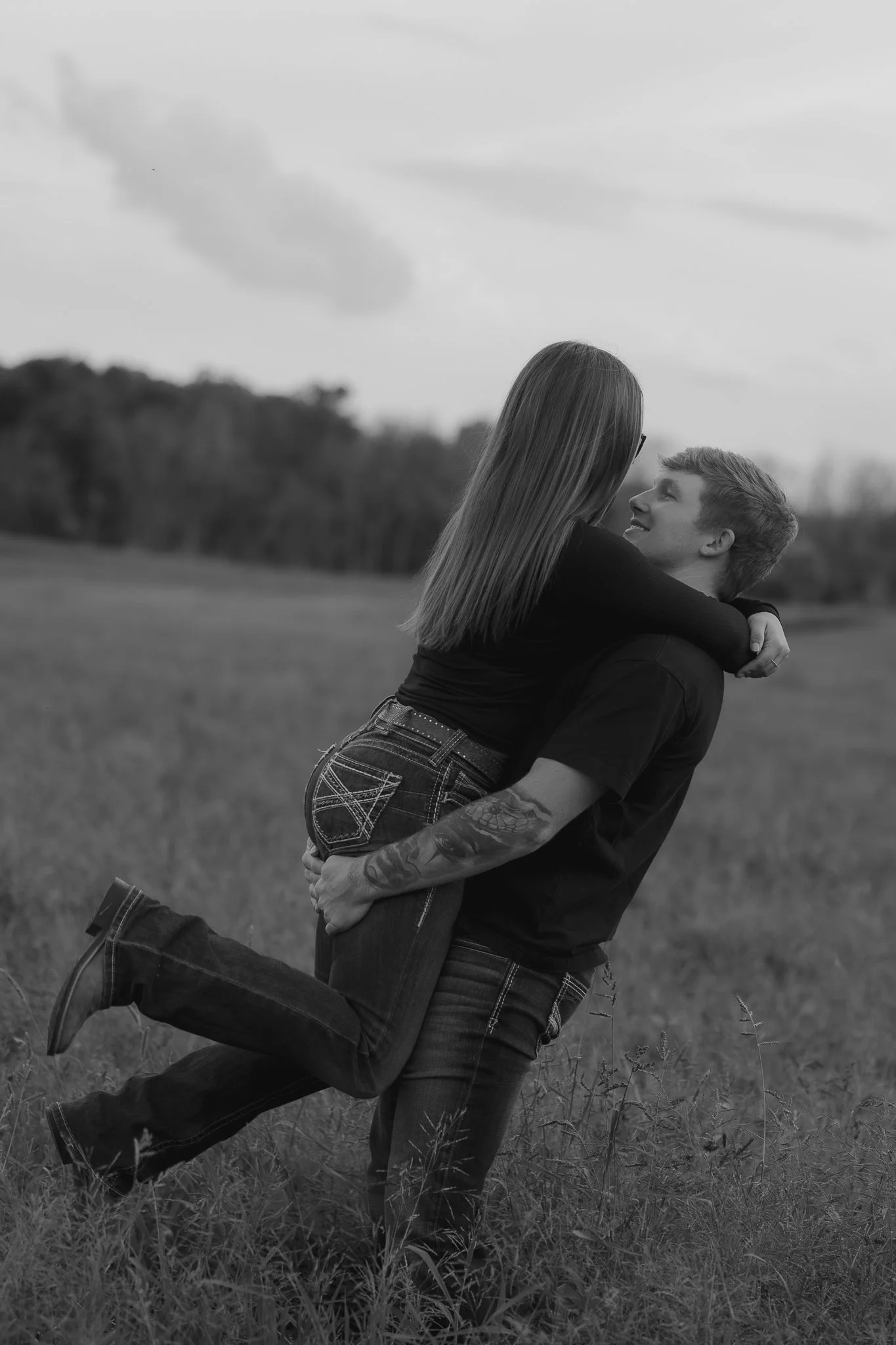 A black and white photo of a couple outdoors. The man is kneeling on one knee, holding the woman around her waist, while the woman is sitting on his lap, with her arms around his shoulders. They are looking at each other and smiling in a field with t