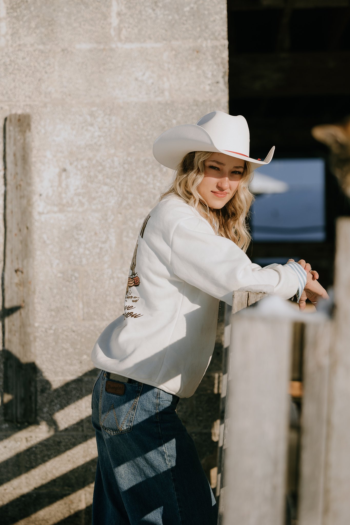 A young woman wearing a white cowboy hat and a white jacket, leaning on a wooden fence, with sunlight casting shadows on her and the background.