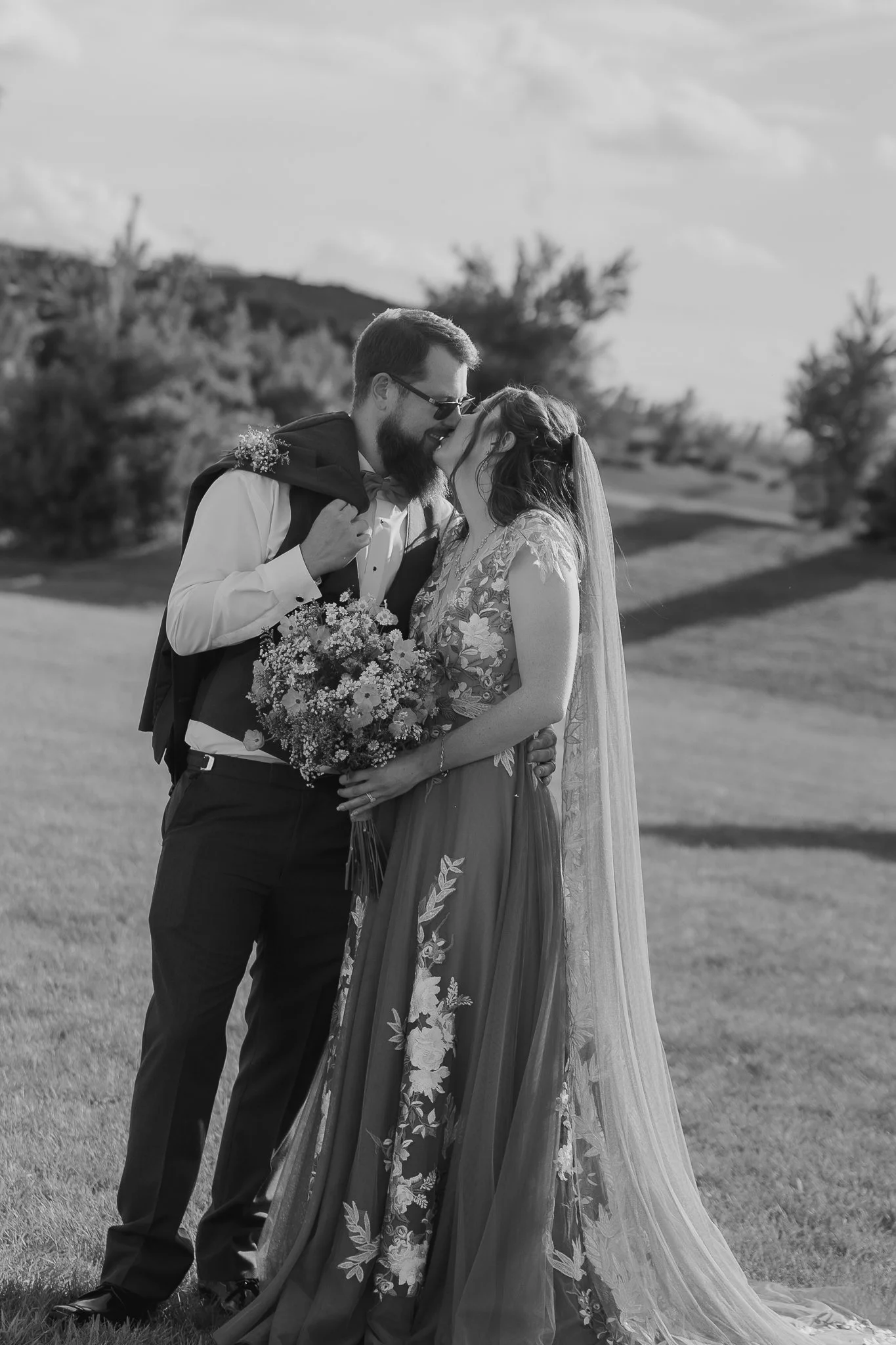 A bride and groom sharing a kiss outdoors; the bride holding a bouquet of flowers; the groom wearing sunglasses and a suit jacket draped over his shoulders.