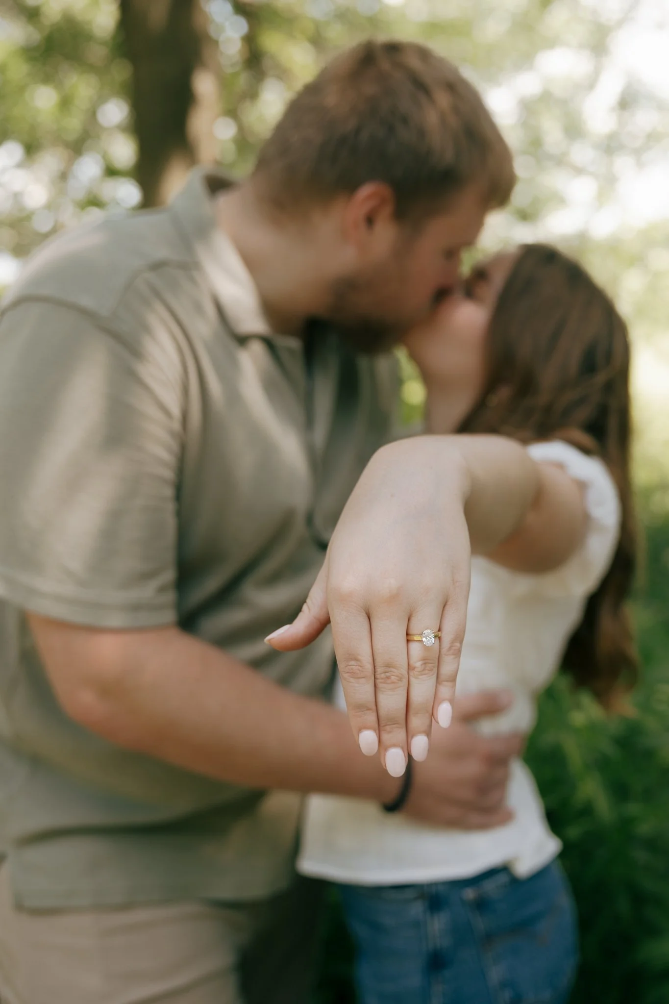 Close-up of a woman's hand showing an engagement ring, with a couple kissing in the background outdoors.