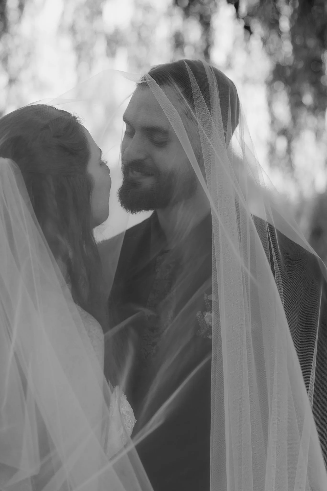 A black-and-white photo of a bride and groom under a wedding veil, close together and smiling lovingly.
