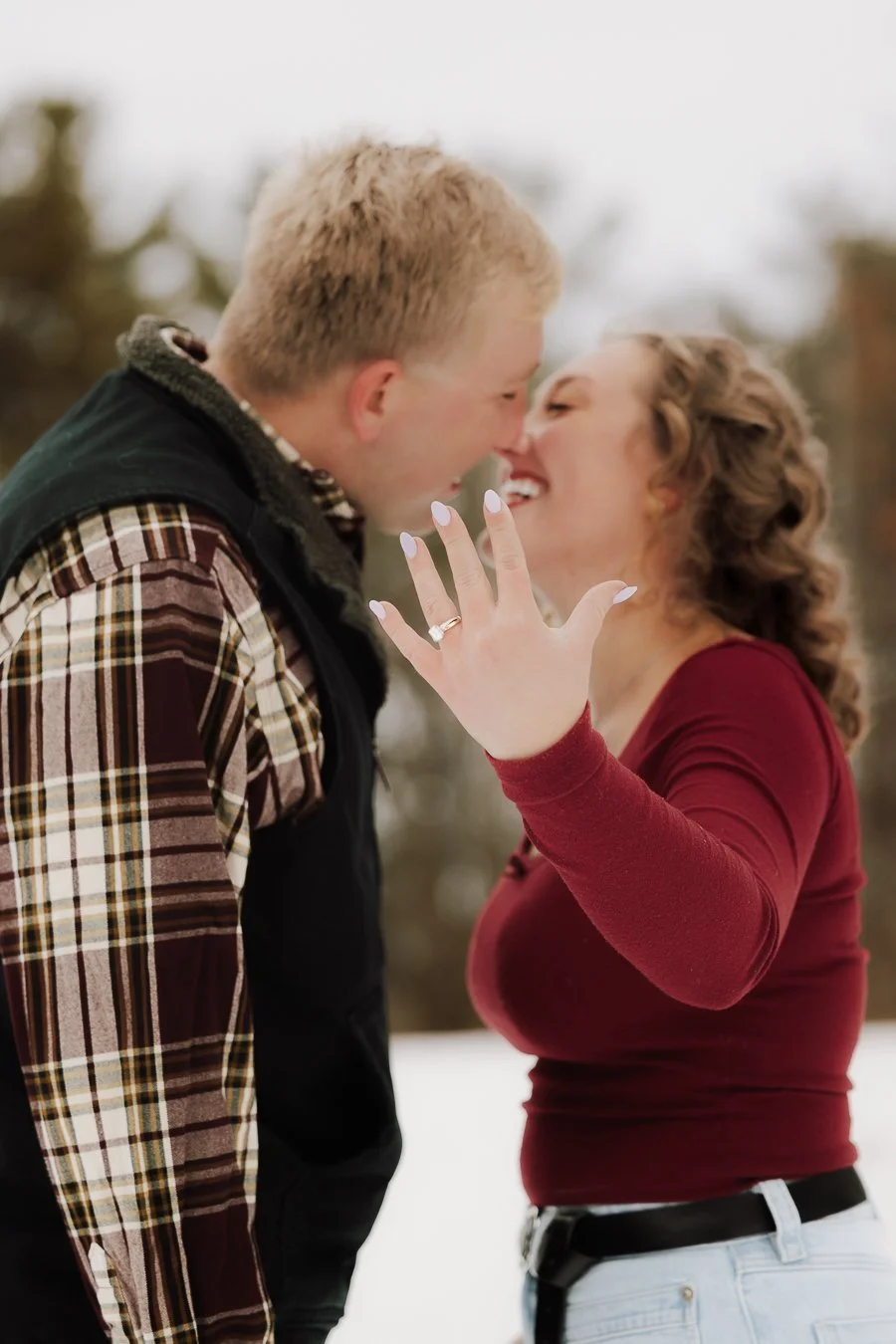 A couple in winter clothing sharing a close, intimate moment outdoors, with the woman showing her engagement ring.