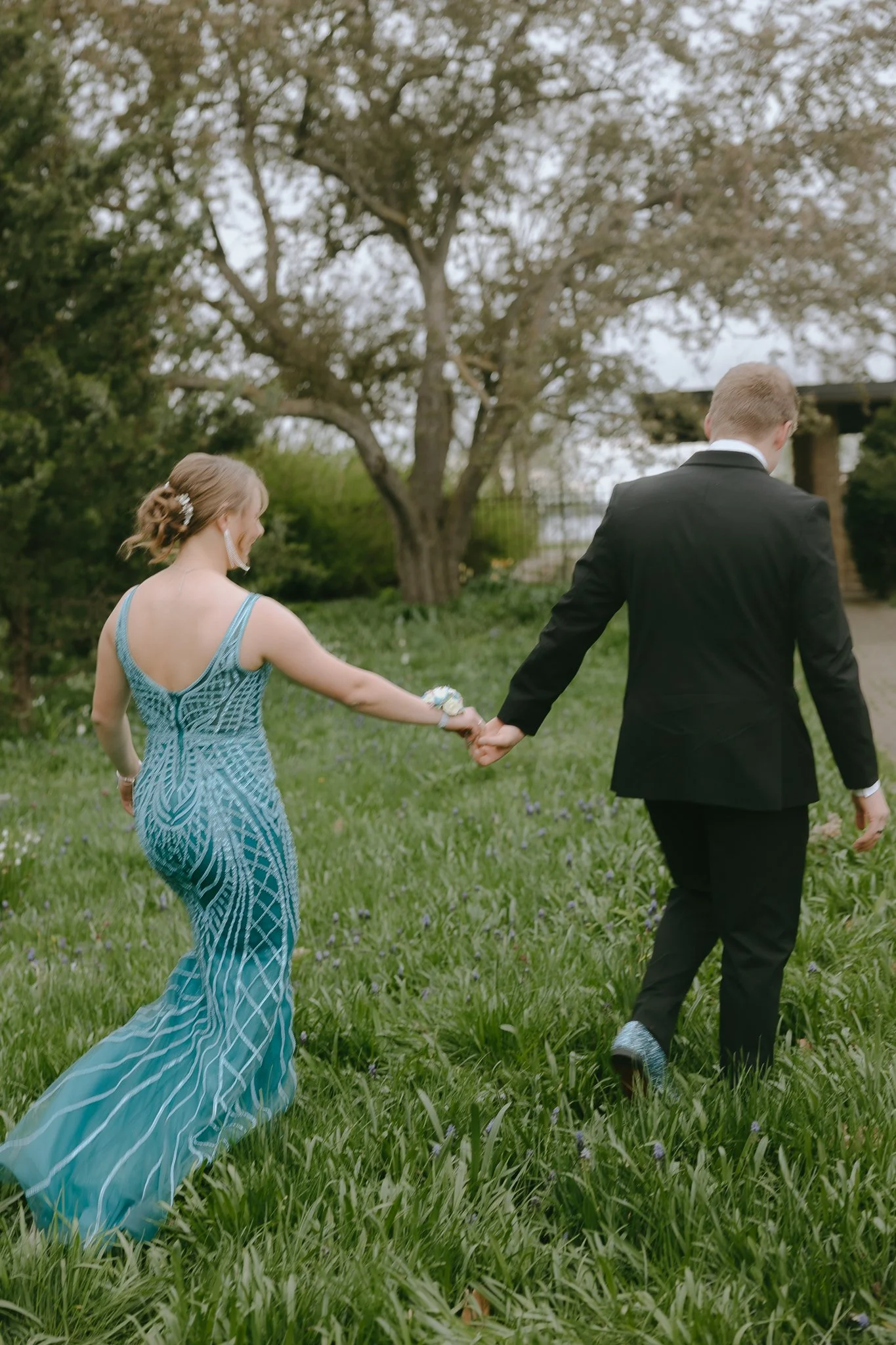 A young couple in formal attire holding hands and walking through a grassy outdoor area with trees in the background.